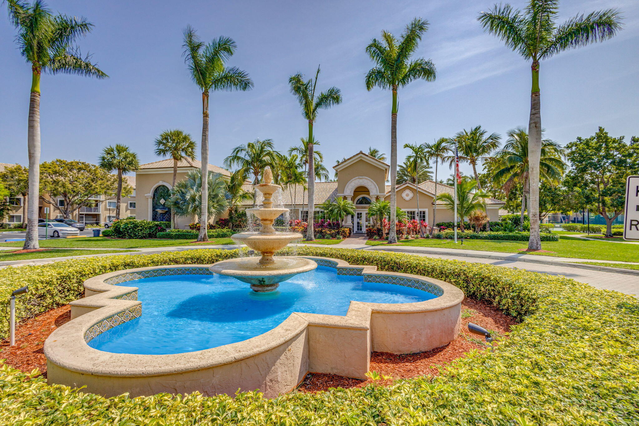 Tiered stone fountain centered within a manicured landscape featuring mature palm trees, sculpted hedges, and concrete walkways