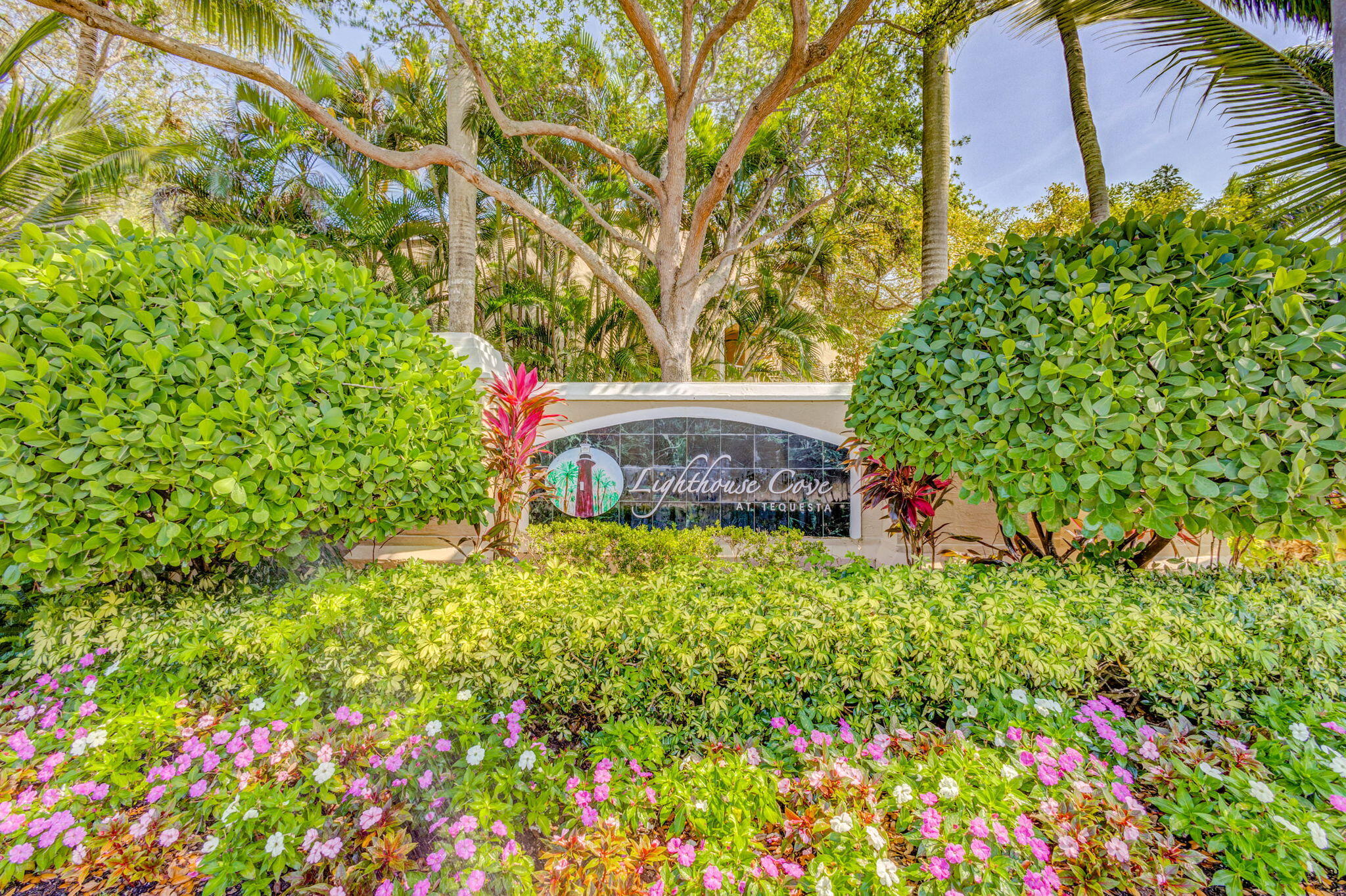Community monument sign featuring decorative stone-finish masonry, mature tropical landscaping, dense shrubbery, and flowering perennials