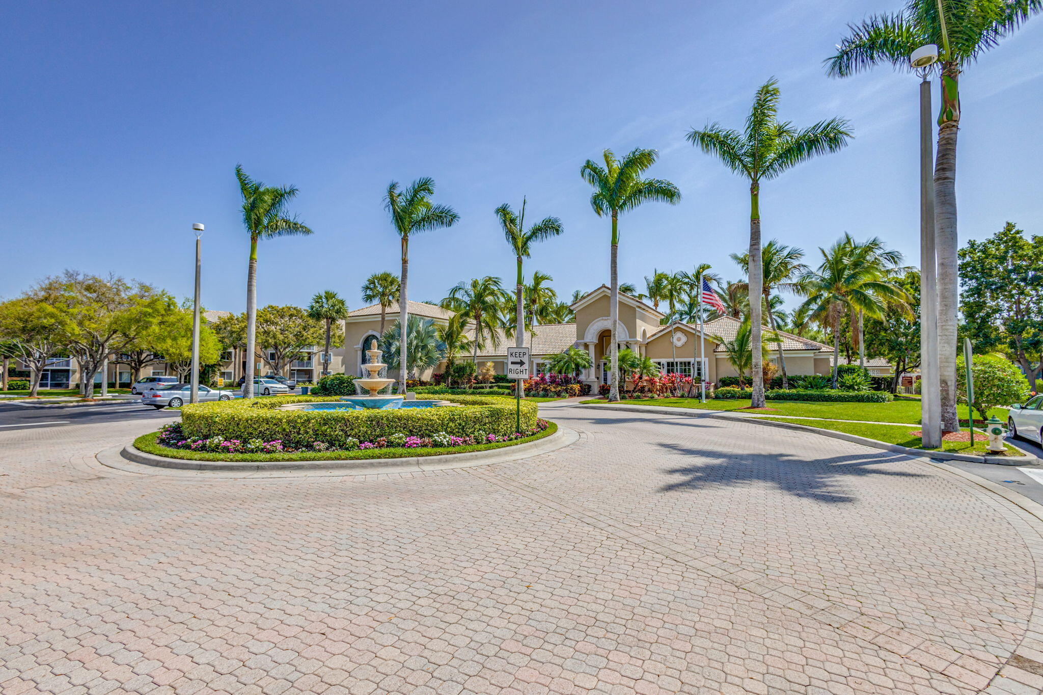 Paved circular drive featuring a central stone-style tiered fountain, manicured perimeter landscaping, and established palm trees