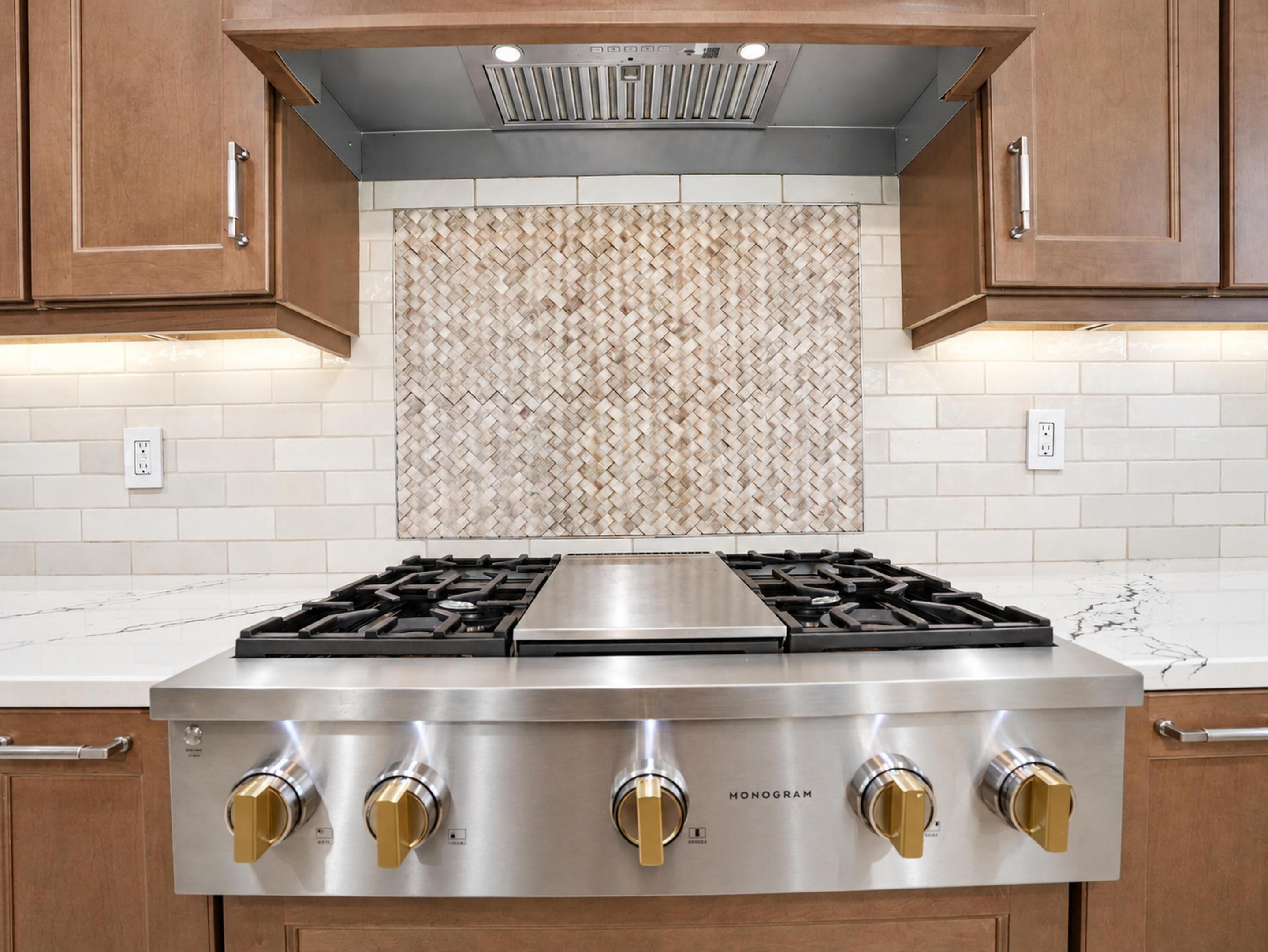 Professional-grade stainless steel gas range with integrated griddle, custom tile backsplash with herringbone pattern insert, stone-finish countertops, and under-cabinet task lighting