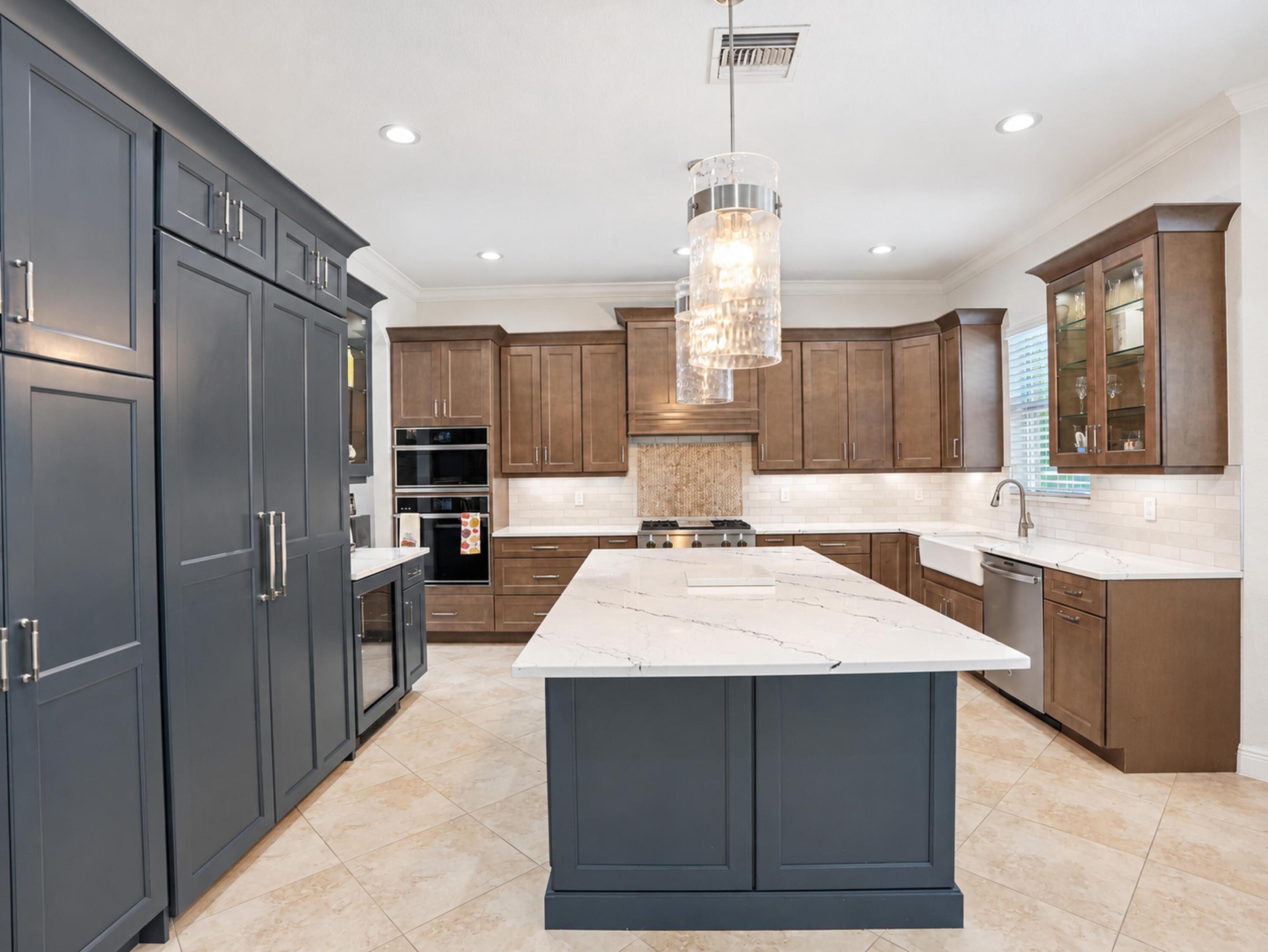 Kitchen featuring an oversized central island with stone-finish countertops, custom two-toned cabinetry, dual built-in wall ovens, porcelain farmhouse sink, and polished glass-paneled pendant lighting