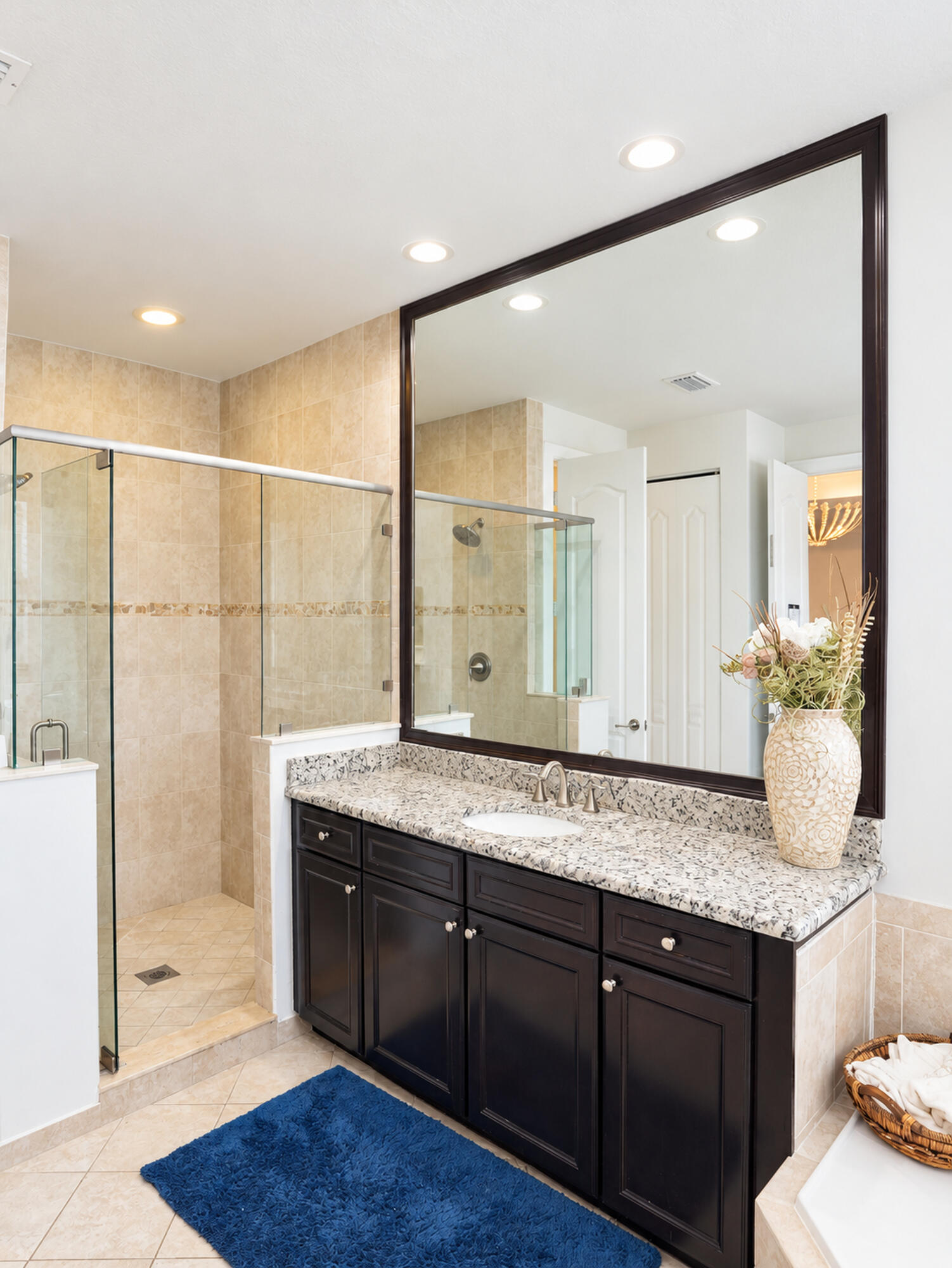 Bathroom vanity with dark cabinetry, stone-surface countertop, oval undermount sink, large framed mirror, and tiled walk-in shower with glass enclosure