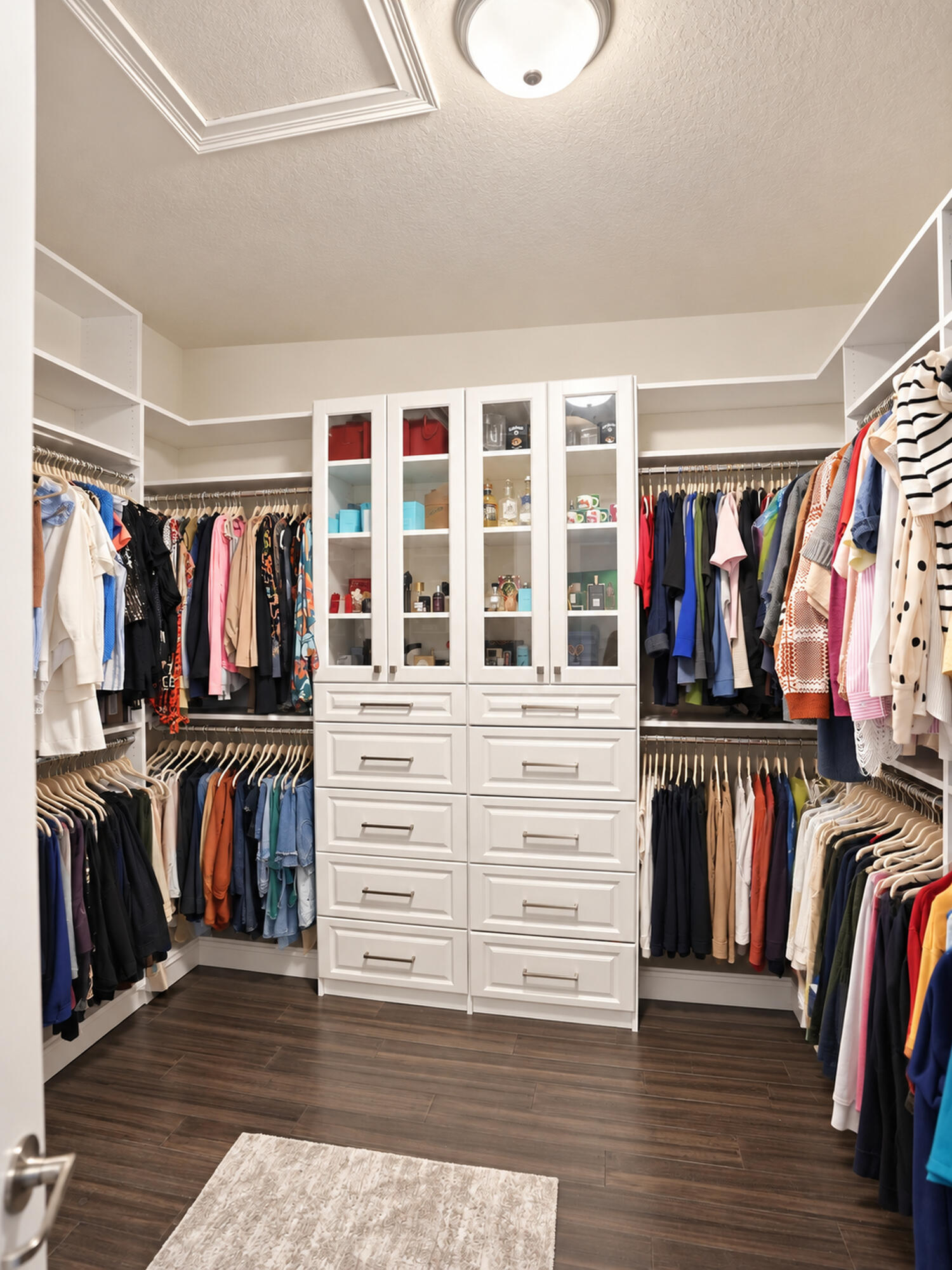 Walk-in closet featuring white built-in cabinetry with glass-front display uppers, stacked storage drawers, wood-finish flooring, and multi-tier hanging rods