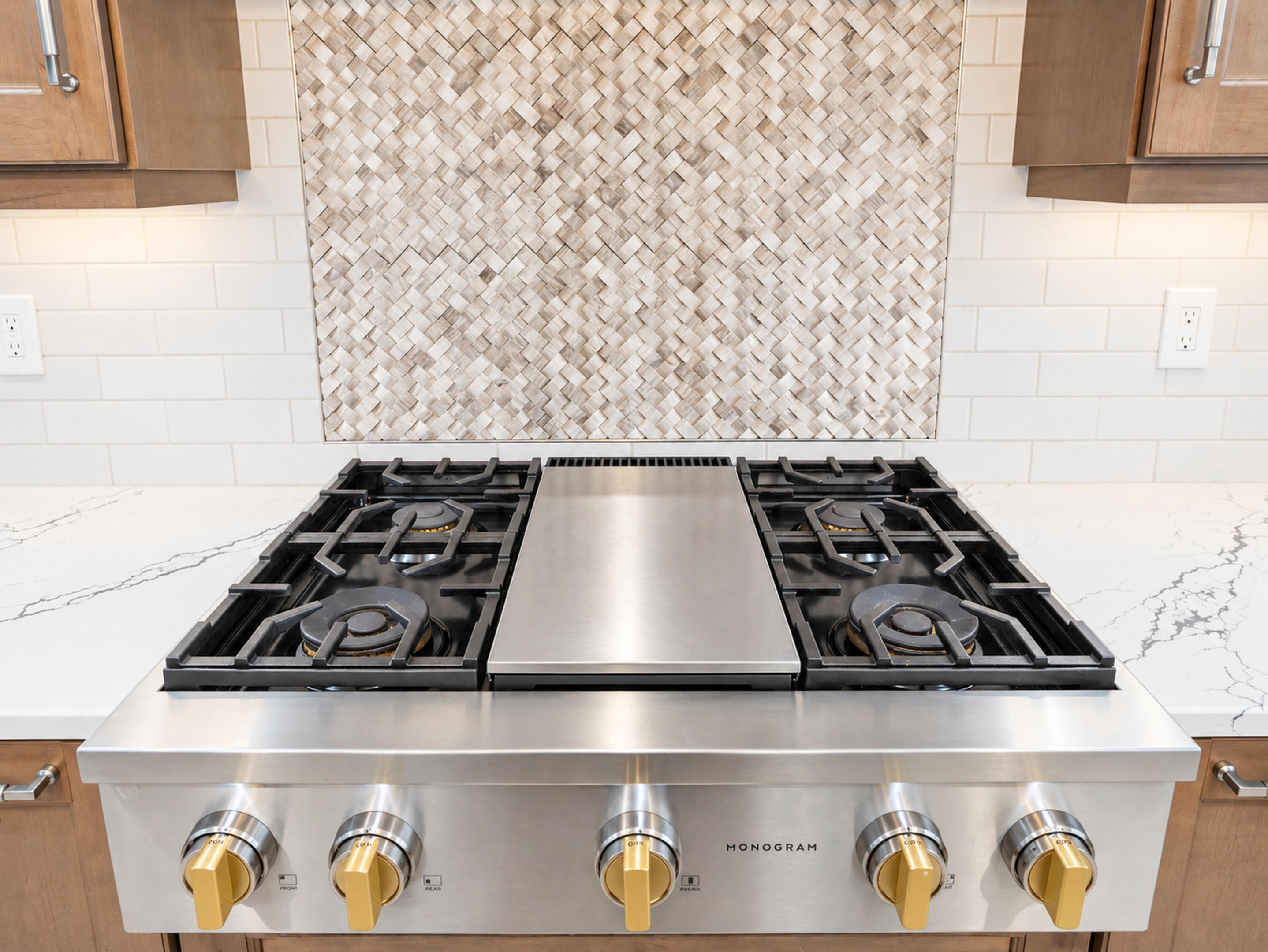 Stainless steel professional-grade gas range with central griddle, light-toned stone-finish countertops, custom herringbone-patterned tile backsplash, and wood-finish cabinetry