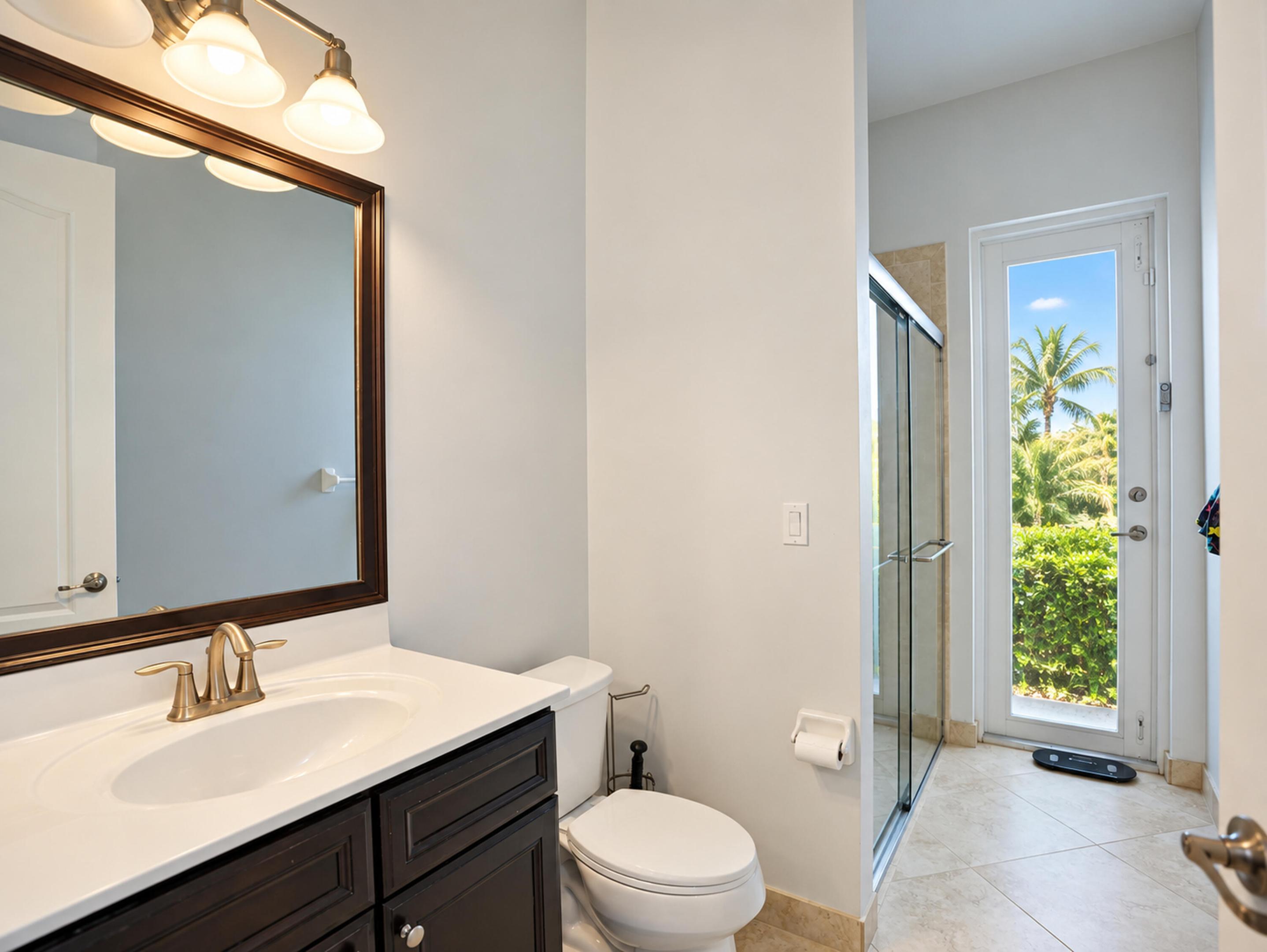 Dark wood-finish vanity with a white countertop and oval undermount sink, framed mirror with integrated vanity lighting, glass-enclosed shower, and full-glass door exterior access