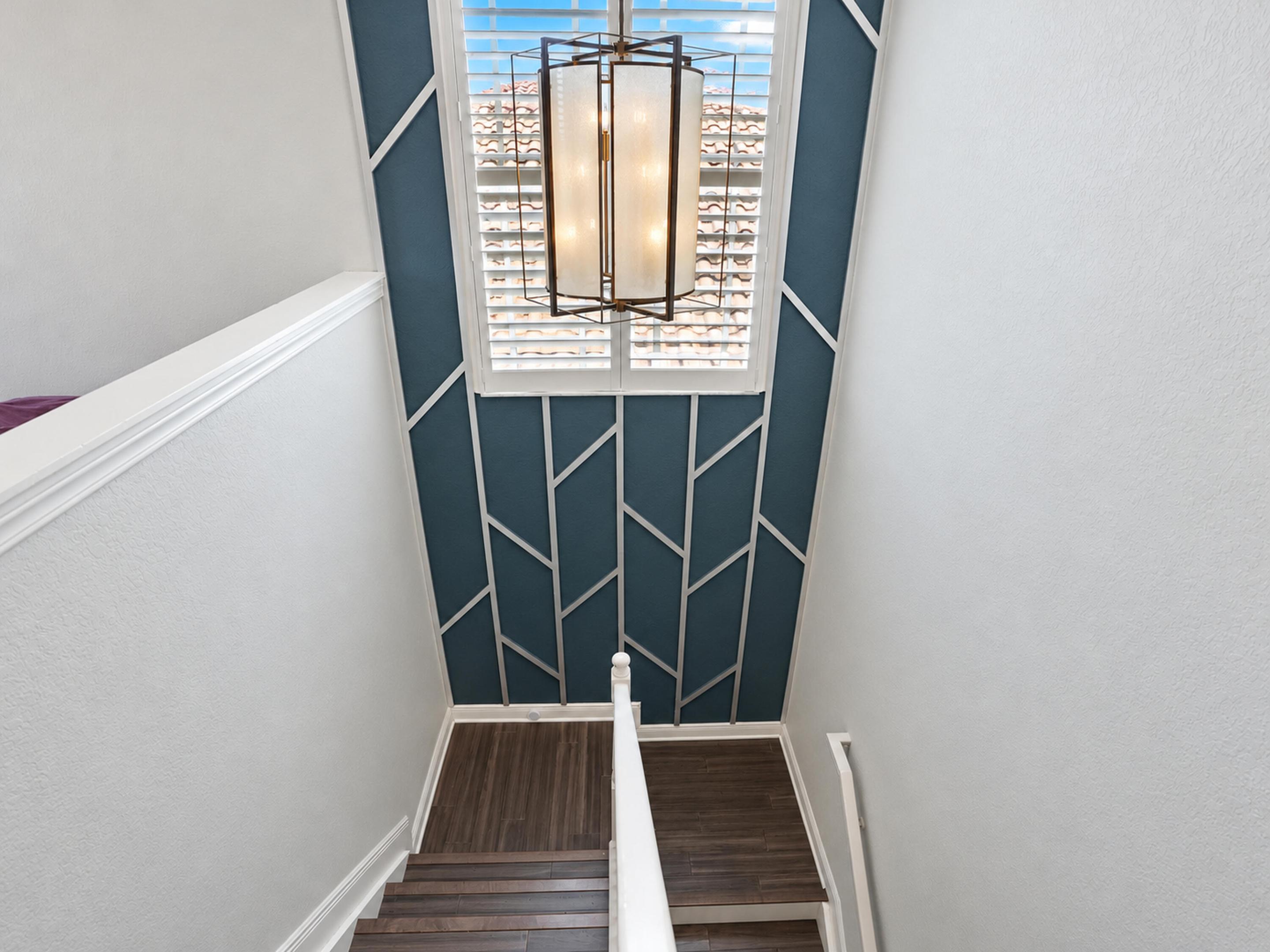 Staircase featuring wood-finish treads, geometric accent wall molding, tiered metal pendant lighting, and plantation shutters