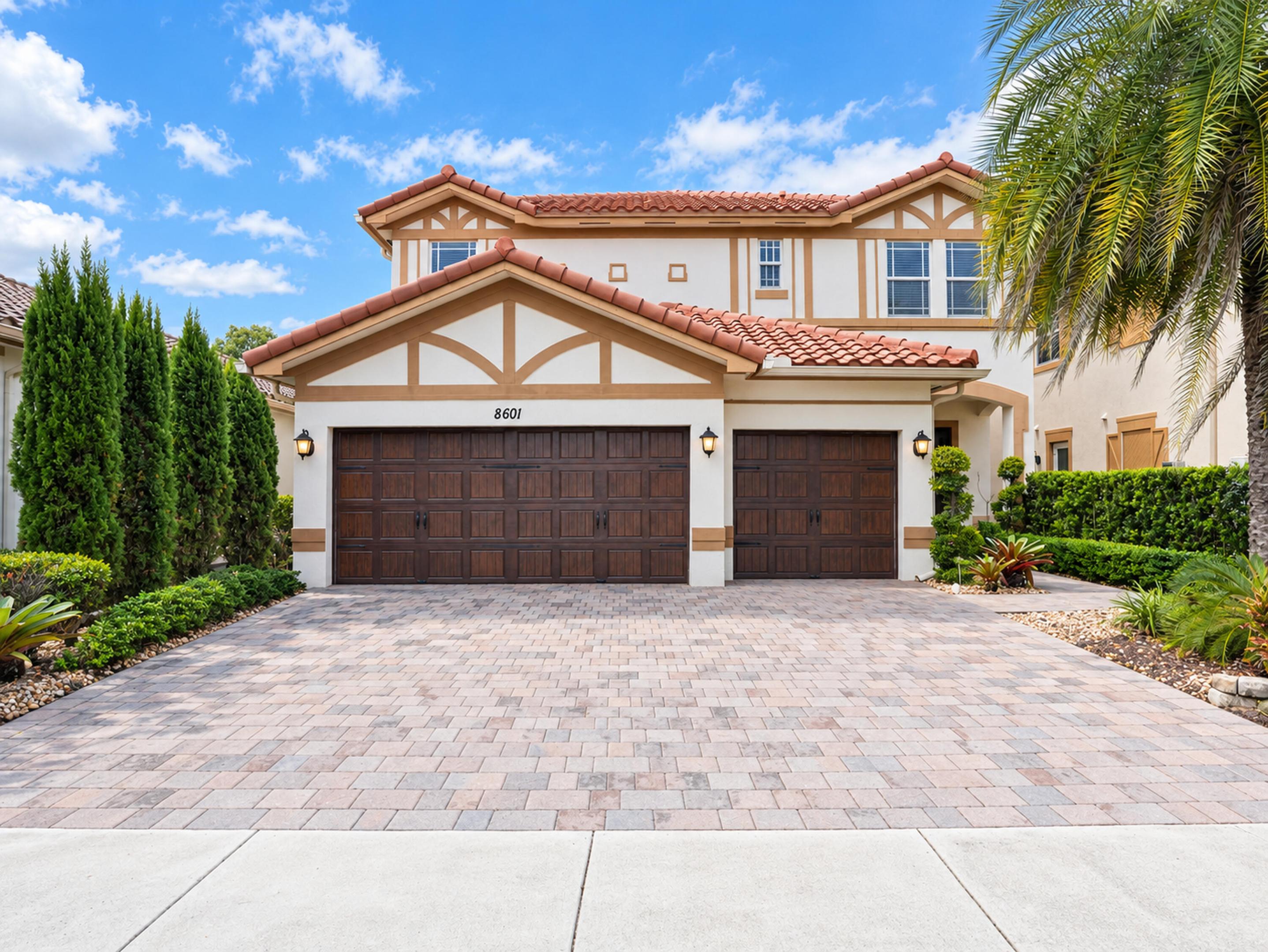 Two-story exterior featuring a clay tile roof, stucco facade with decorative timbering, paver driveway, and a three-car garage with wood-finish doors