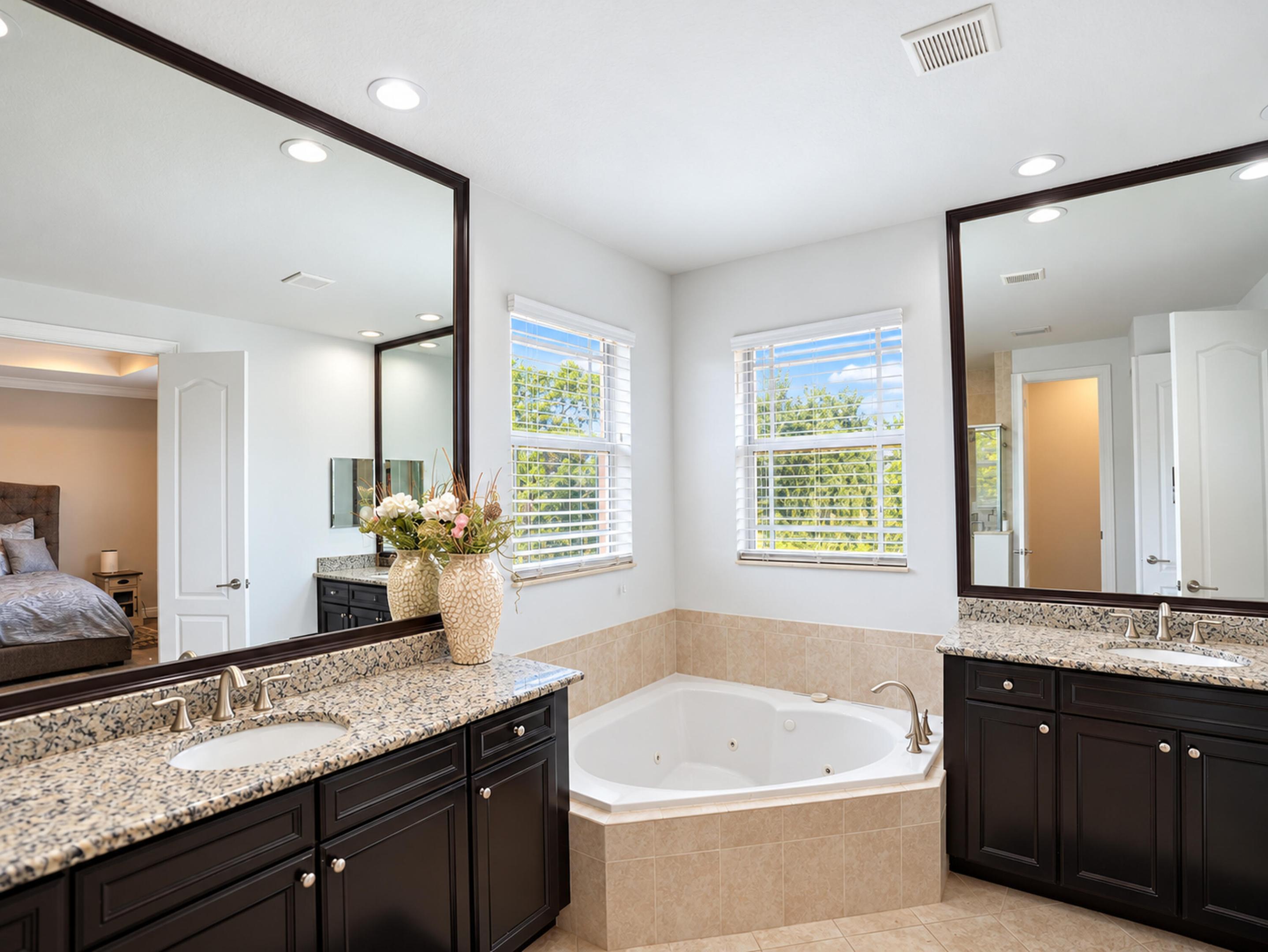 Dual vanities with dark-finish cabinetry, stone-style countertops, integrated undermount sinks, and large-format framed mirrors, featuring a soaking jetted tub set in a tile-surround platform