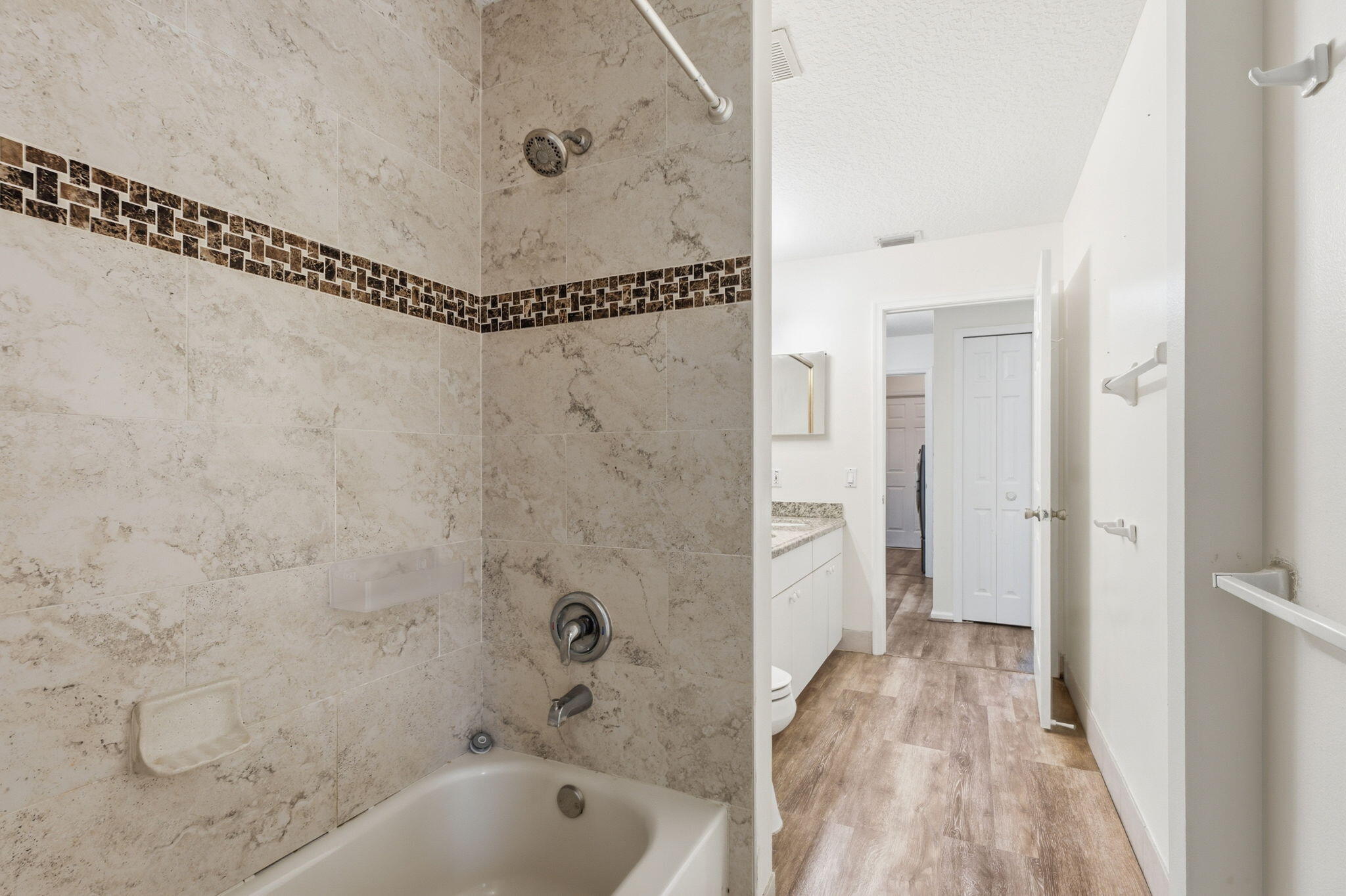 Shower area featuring stone-finish wall tile with mosaic inlay, a chrome shower head and handle, white cabinetry, and wood-finish flooring