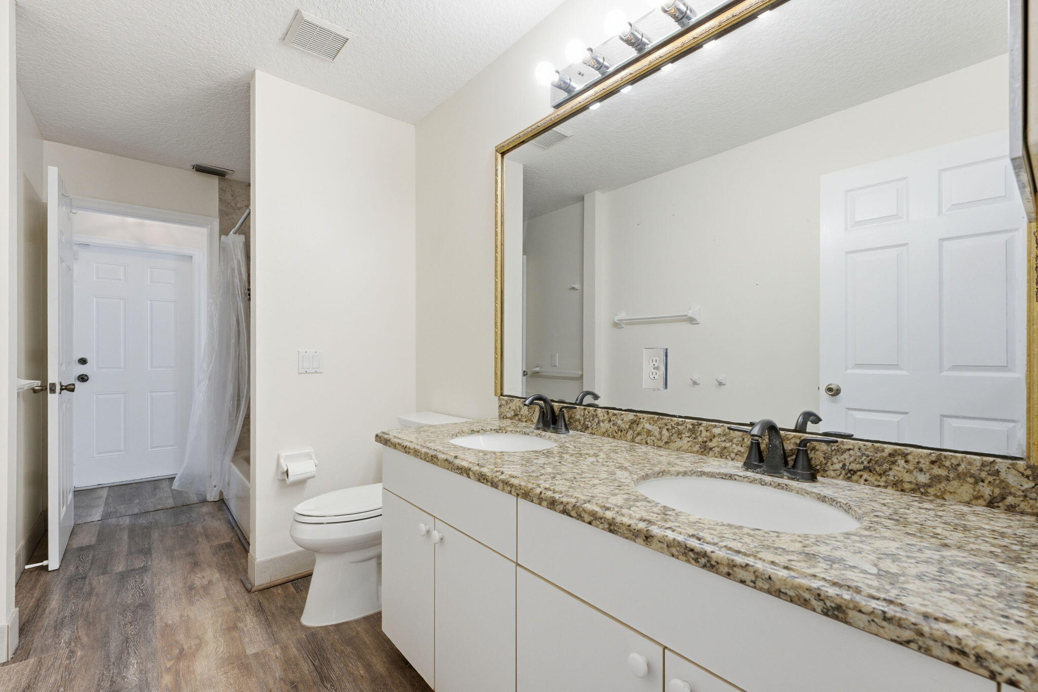 Dual-sink vanity with stone-finish countertop, undermount sinks, bronze-tone fixtures, wood-finish flooring, and large wall-mounted mirror