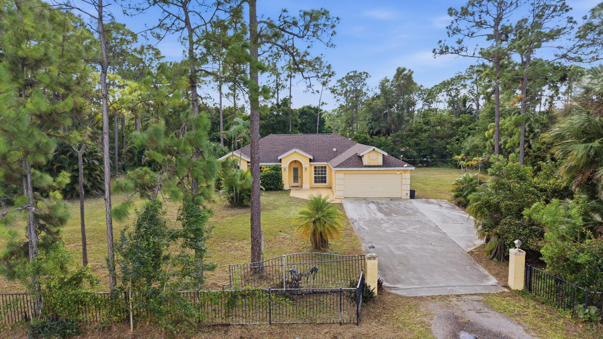 Single-story residence featuring a hipped roof, attached garage, concrete driveway, and perimeter metal fencing set among mature pine trees