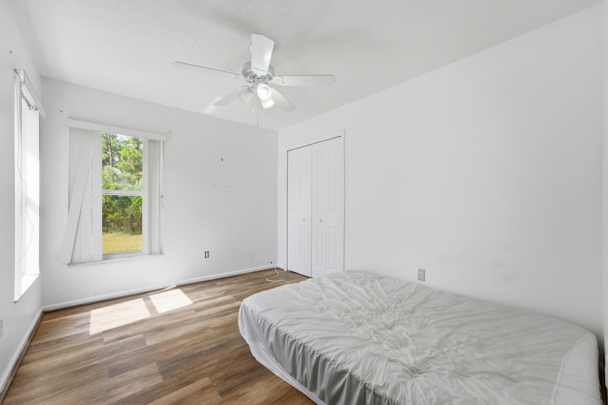 Neutral-toned interior space featuring wood-finish flooring, a white ceiling fan with integrated lighting, dual-pane windows with vertical blinds, and a double-door reach-in closet