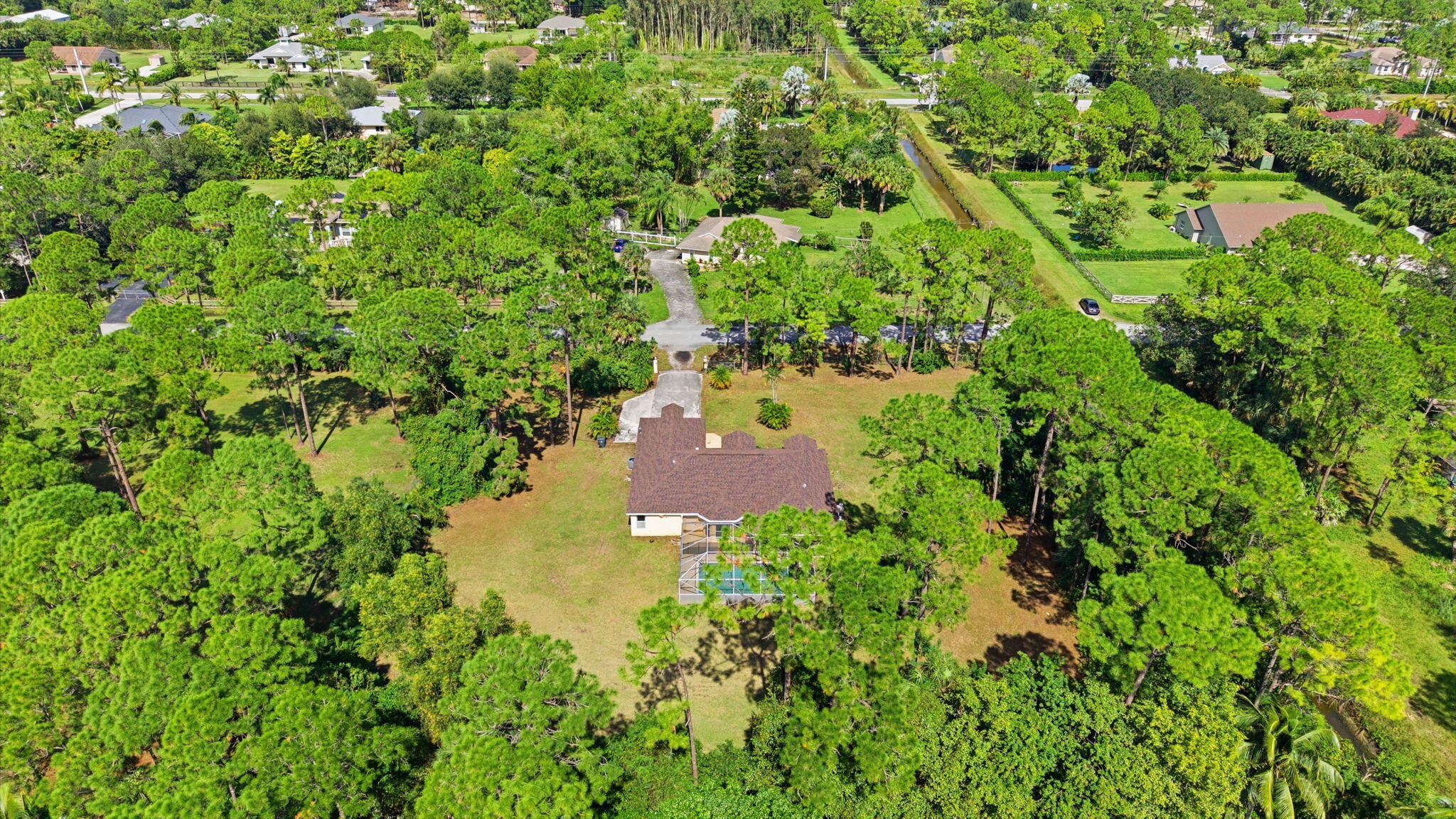 Aerial perspective featuring a single-story residence situated on a spacious lot, expansive grass lawn, mature trees, and a rectangular swimming pool with an enclosure