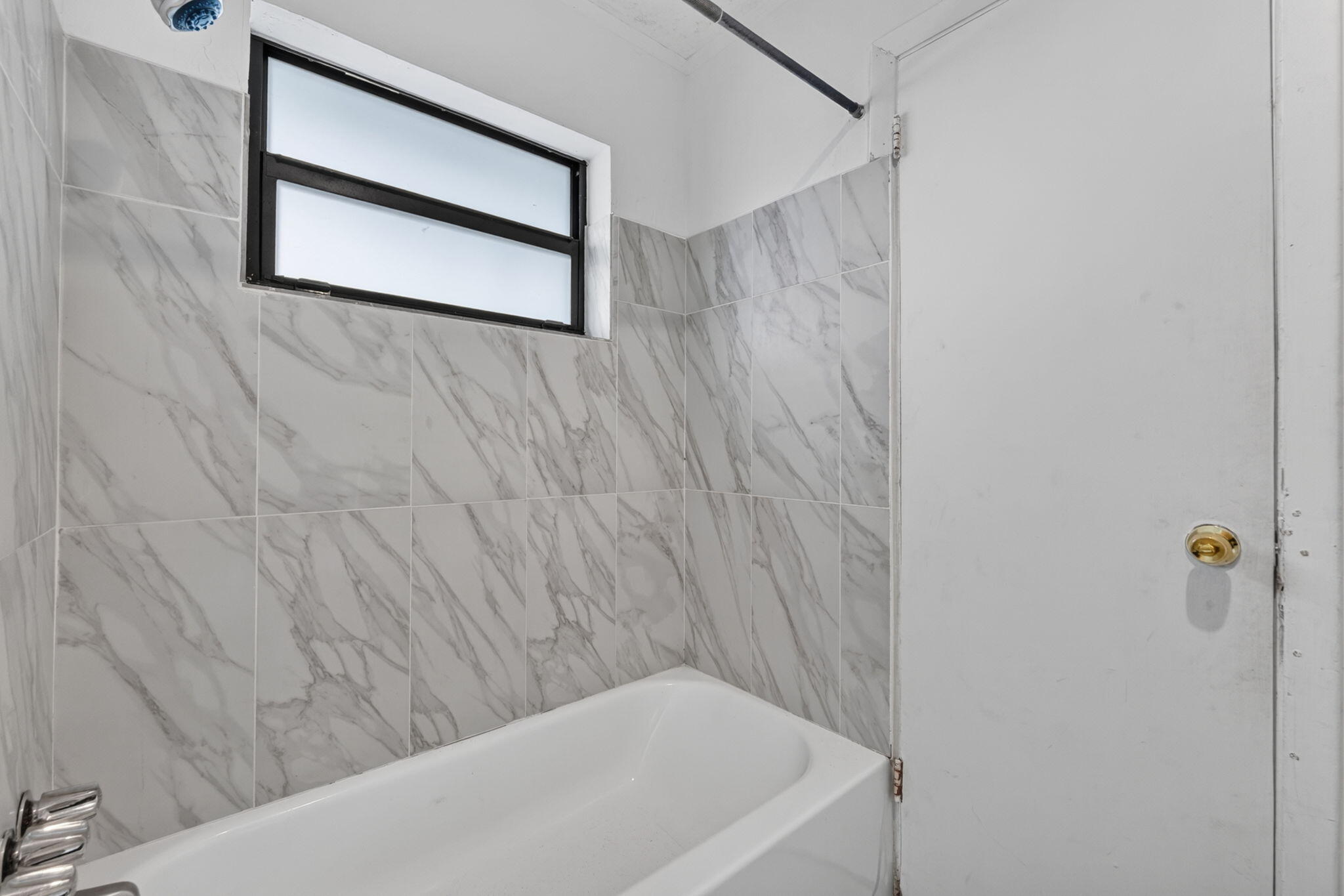 Bathtub featuring light-toned stone-patterned wall tile, integrated chrome hardware, and a single horizontal frosted-glass window