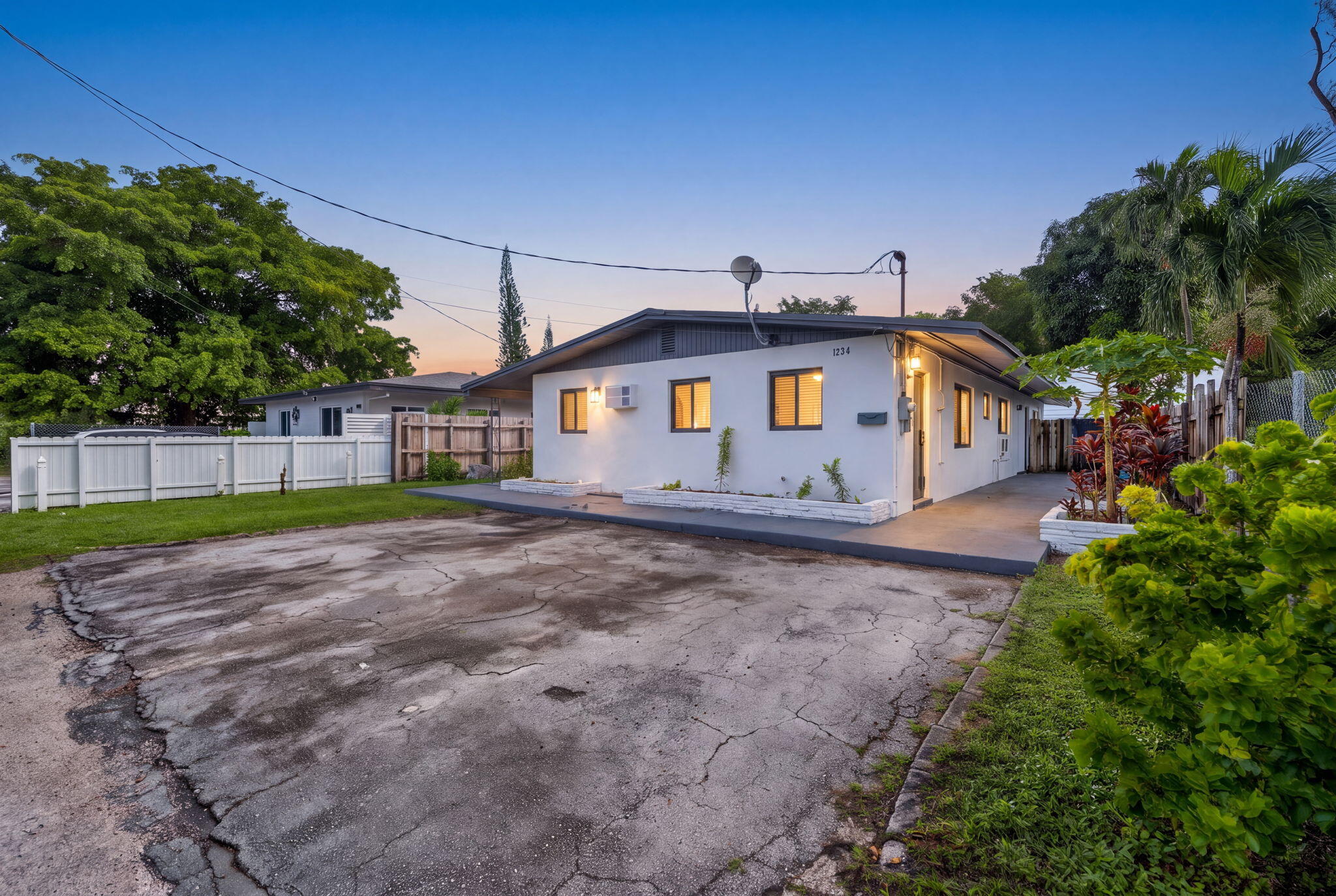 Single-story exterior featuring white stucco siding, a grey-toned roofline, paved driveway, and perimeter white vinyl fencing