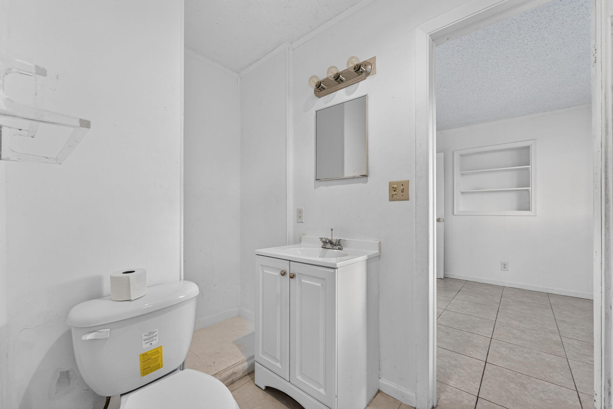 White vanity cabinet with an integrated sink and chrome faucet, wall-mounted light fixture, square mirror, and built-in wall shelving