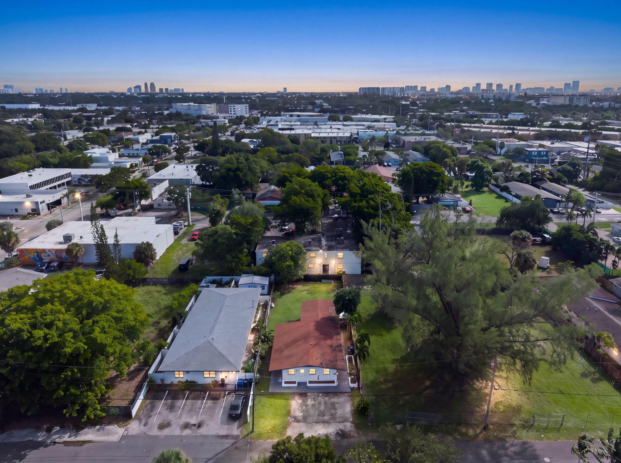 Aerial perspective of a property featuring expansive green space, mature canopy trees, paved off-street parking, and a distant city skyline