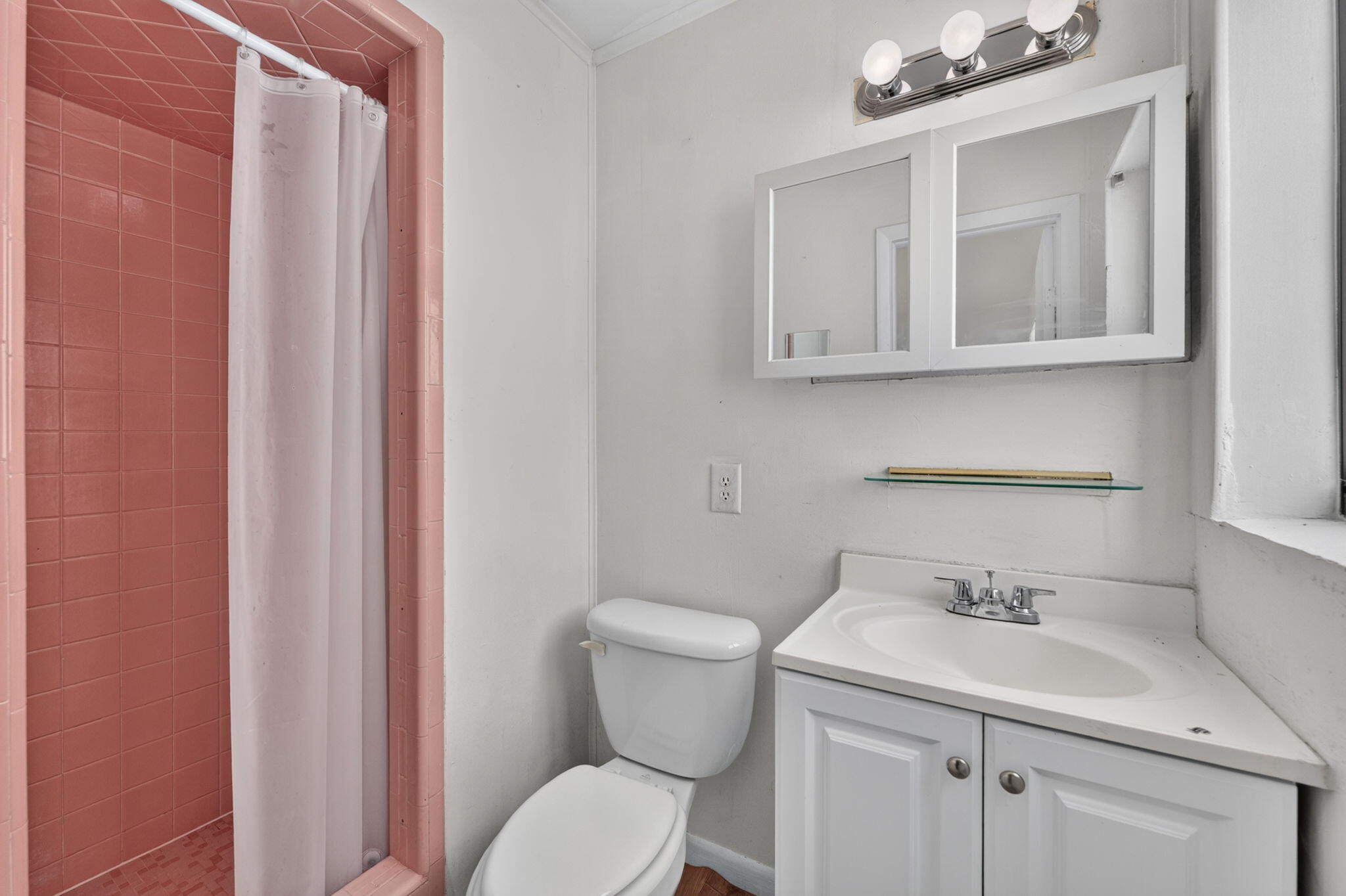 Bathroom featuring a shower stall with pink tile surround, white vanity cabinet with a integrated sink, double-door medicine cabinet, and three-bulb vanity light fixture