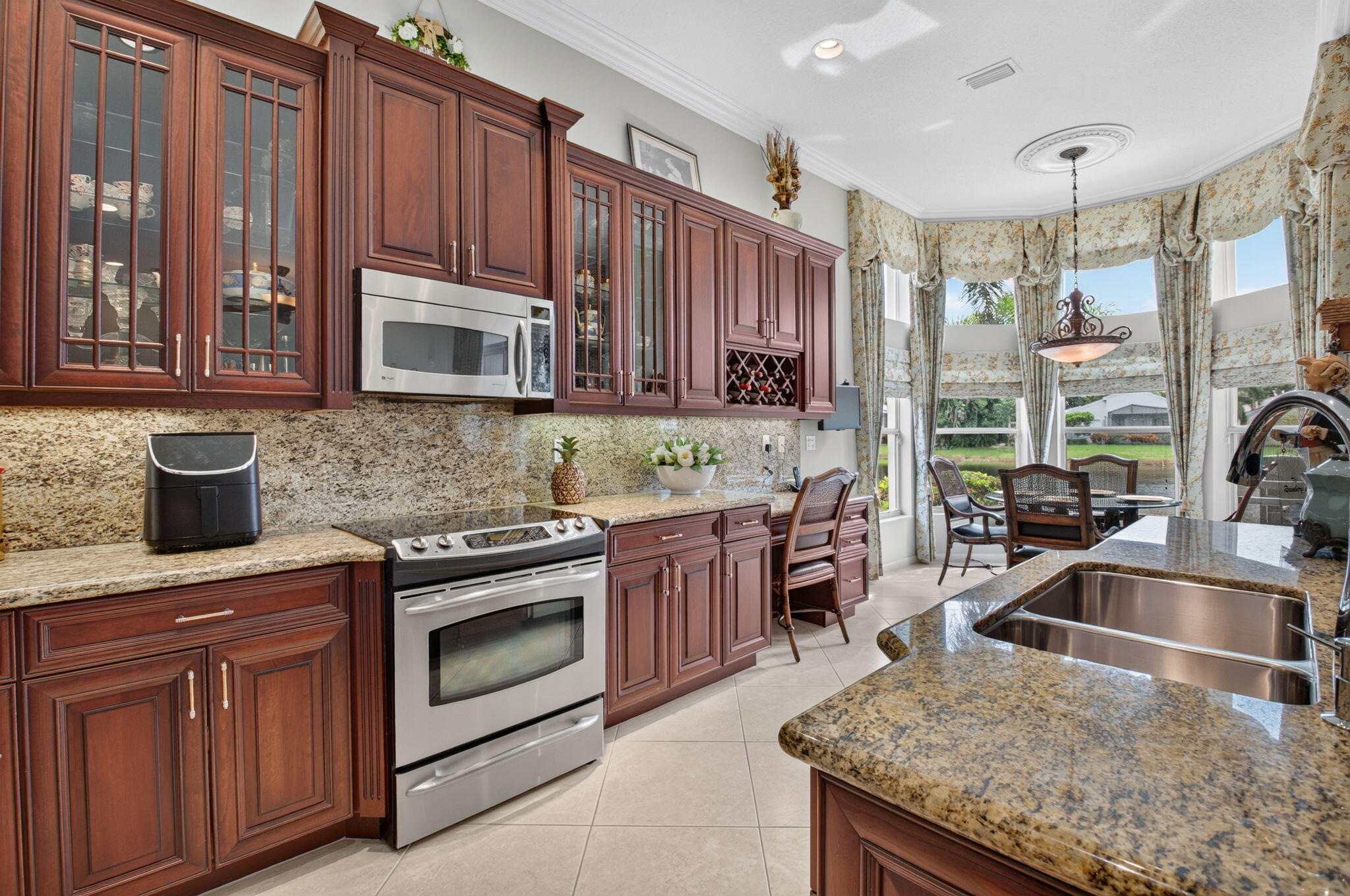 Kitchen featuring raised-panel cherry wood cabinetry, stone countertops, integrated wine rack, stainless steel appliance suite, and a multi-paneled bay window with crown molding