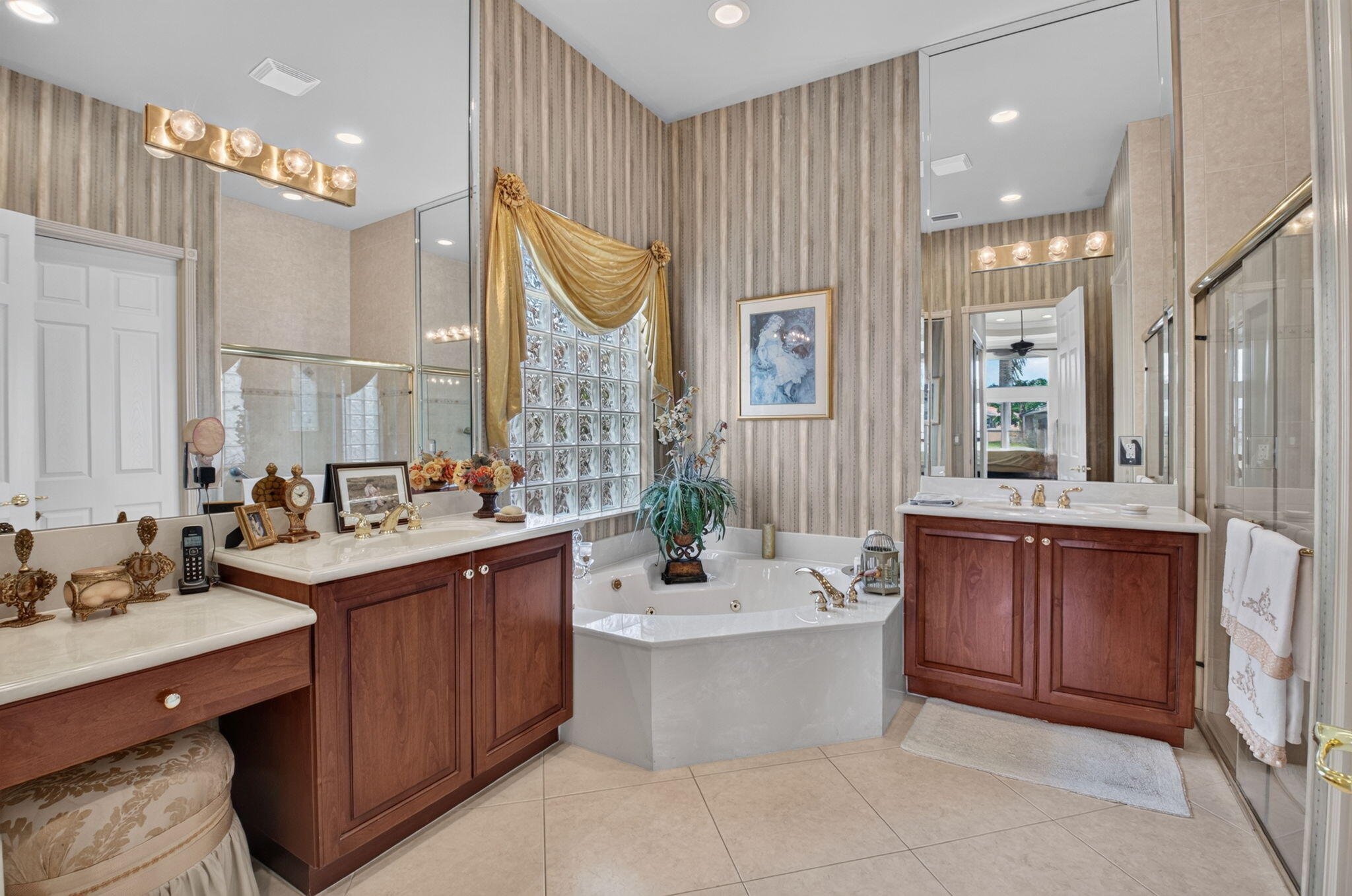 Bathroom featuring dual wood-finish vanities with stone countertops, a corner jetted soaking tub, glass block accent window, and wall-to-wall mirror panels