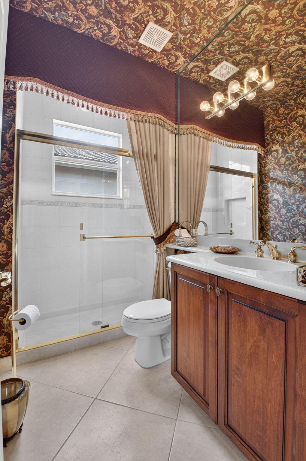 Bathroom featuring wood-finish vanity cabinetry, a rectangular white countertop with an integrated sink, glass-enclosed shower with gold-tone hardware, and large-format tile flooring