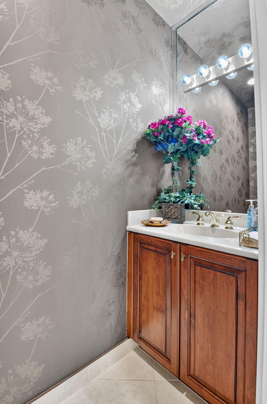 Vanity featuring a stone-composite countertop, undermount sink, brass-finish faucet, and wood-toned cabinetry, accented by botanical-print wall coverings and a multi-bulb vanity light fixture