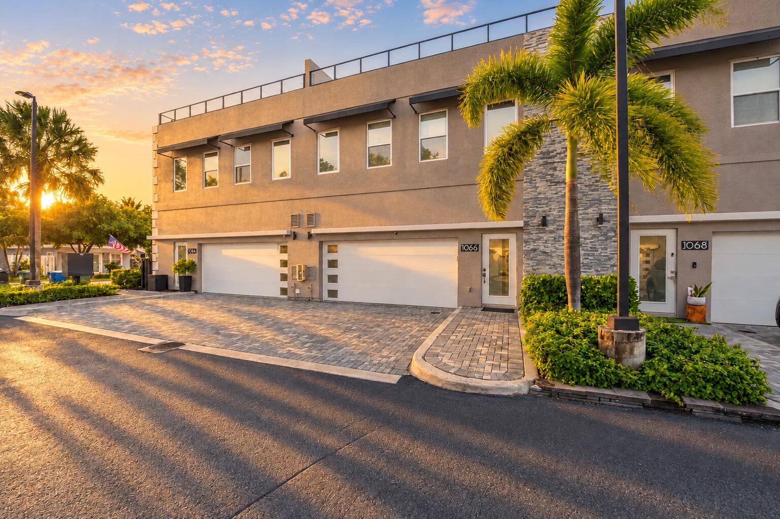 Stucco exterior featuring stacked stone accents, multi-car garage with modern paneled doors, rooftop terrace railing, and paver driveway Stucco exterior featuring stacked stone accents, multi-car garage with modern paneled doors, rooftop terrace railing, and paver driveway