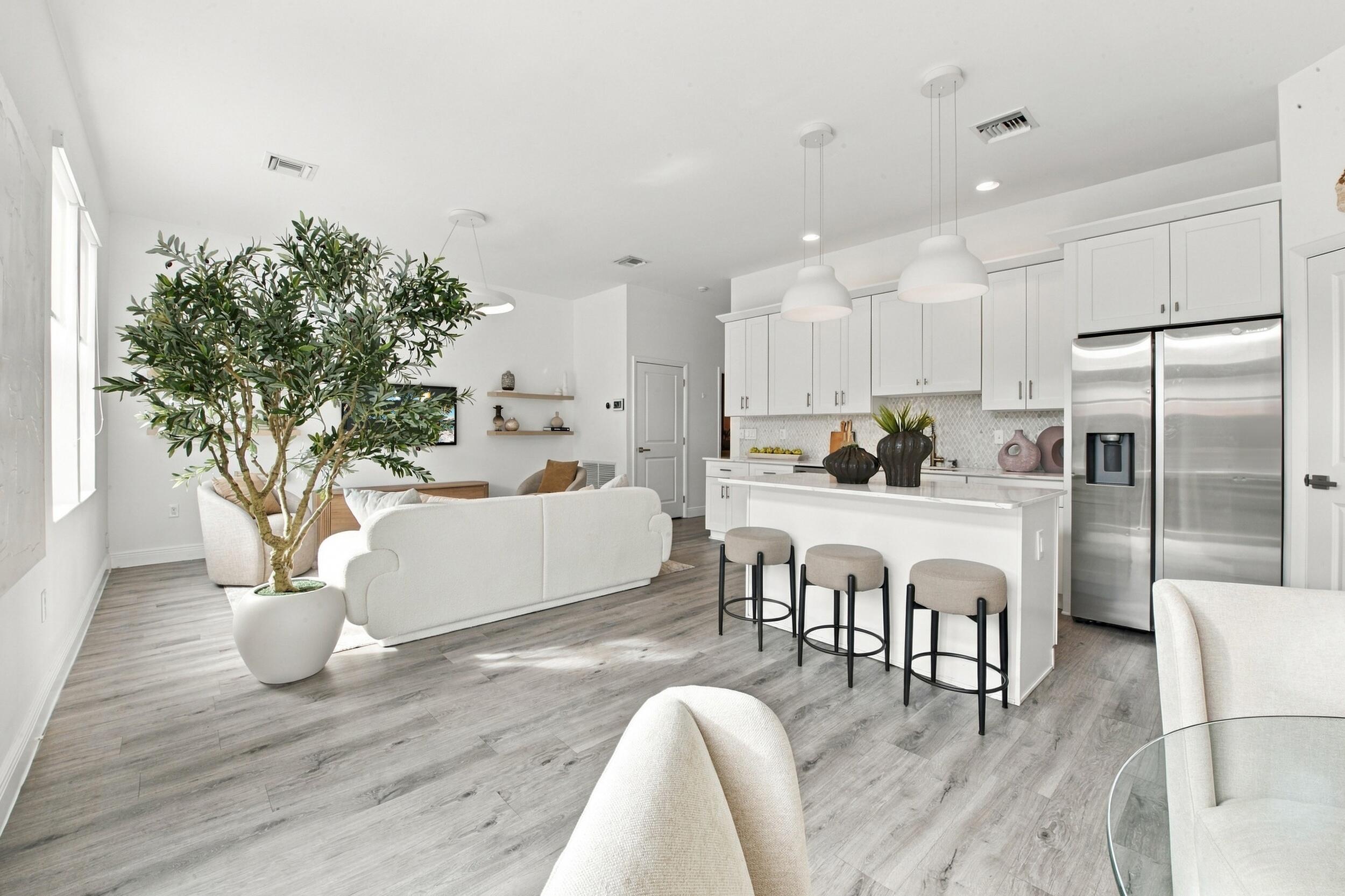 Open-concept interior featuring wood-finish flooring, white shaker cabinetry, stainless steel refrigerator, kitchen island with seating, and pendant lighting Open-concept interior featuring wood-finish flooring, white shaker cabinetry, stainless steel refrigerator, kitchen island with seating, and pendant lighting