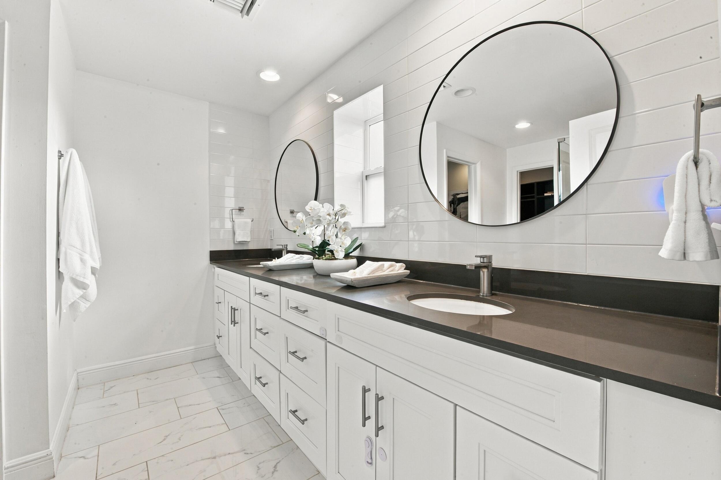 Dual-sink vanity featuring white cabinetry, a solid-surface countertop, rectangular undermount sinks, wall-mounted round mirrors, and tile flooring