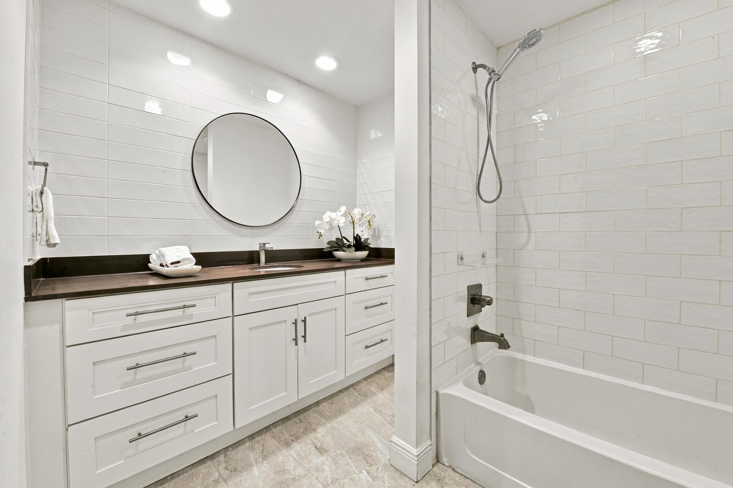 Bathroom featuring a white shaker-style vanity with a dark countertop, round wall-mounted mirror, ceramic subway tile wall cladding, and a tub-shower combination with bronze-finish fixtures