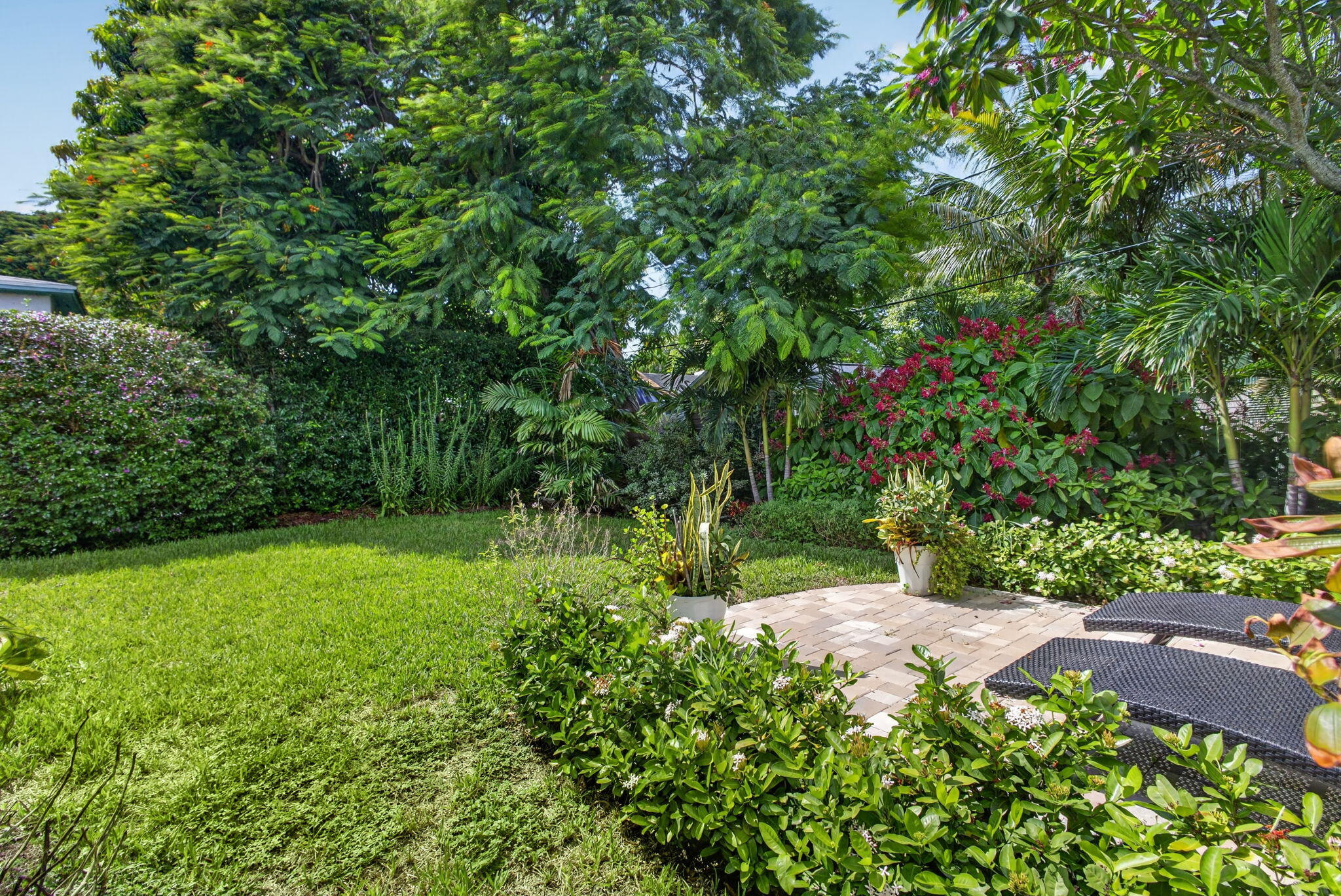 Landscaped backyard featuring a paved stone patio, mature trees, flowering shrubs, and a lawn