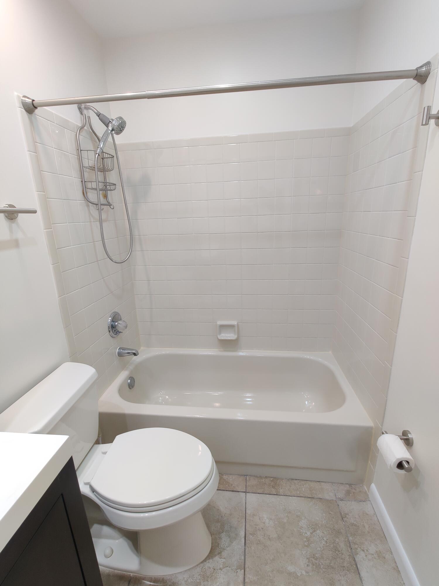 Bathtub and shower combination featuring light-toned wall tile, a handheld showerhead, a neutral vanity, and stone-finish flooring