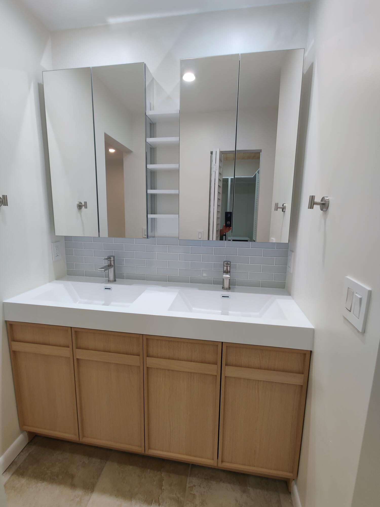 Double vanity featuring a white integrated countertop, light wood-finish cabinetry, a glass-tiled backsplash, dual chrome faucets, and mirrored medicine cabinets with integrated shelving