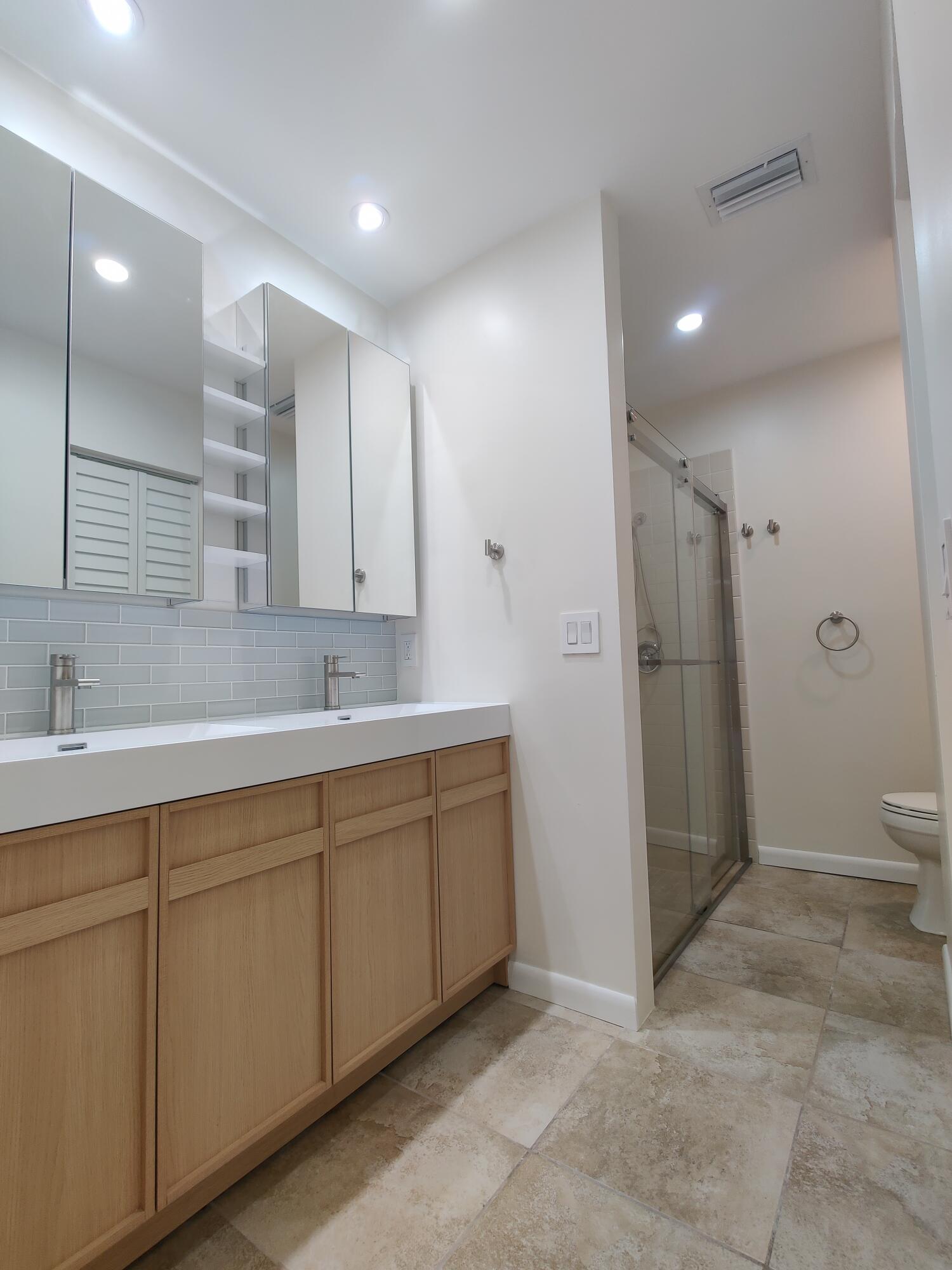 Dual-sink vanity with light wood cabinetry, integrated white countertops, recessed lighting, and a glass-enclosed shower with tile flooring