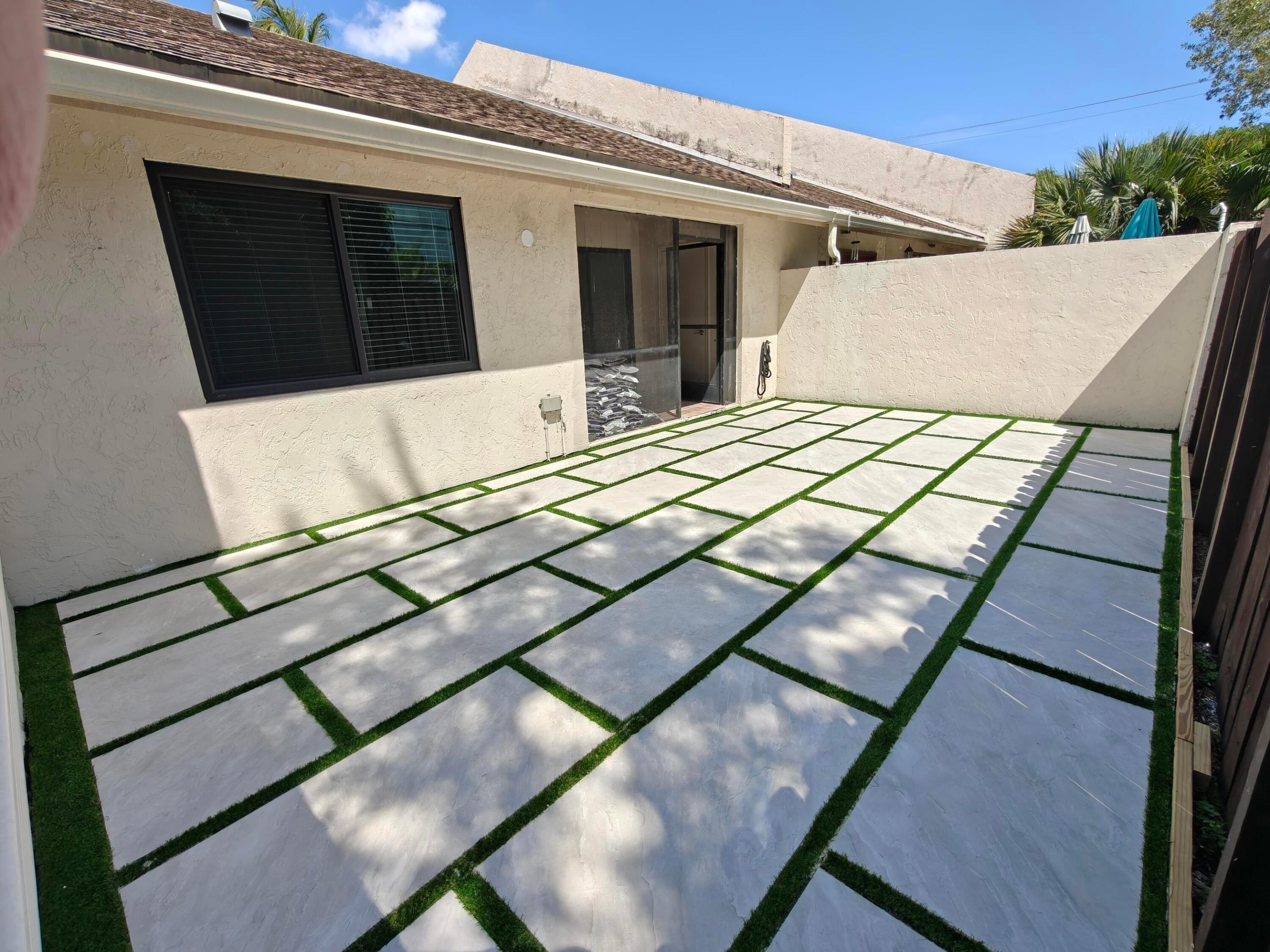 Private rear courtyard featuring large-format concrete pavers with artificial turf inlay, enclosed by a stucco perimeter wall and a wood-slat fence