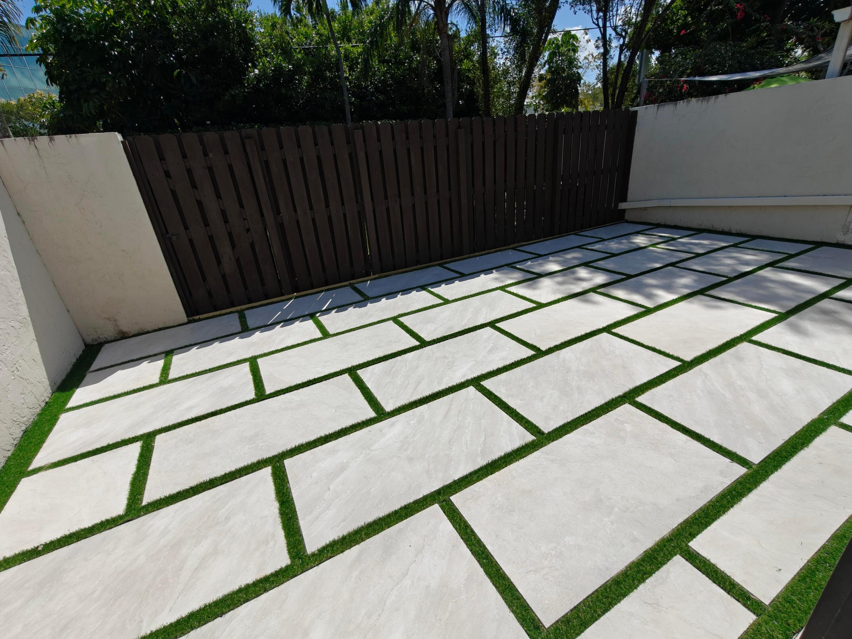 Outdoor patio featuring oversized light-toned stone pavers, contrasting synthetic turf inlay, and wood-finish privacy fencing