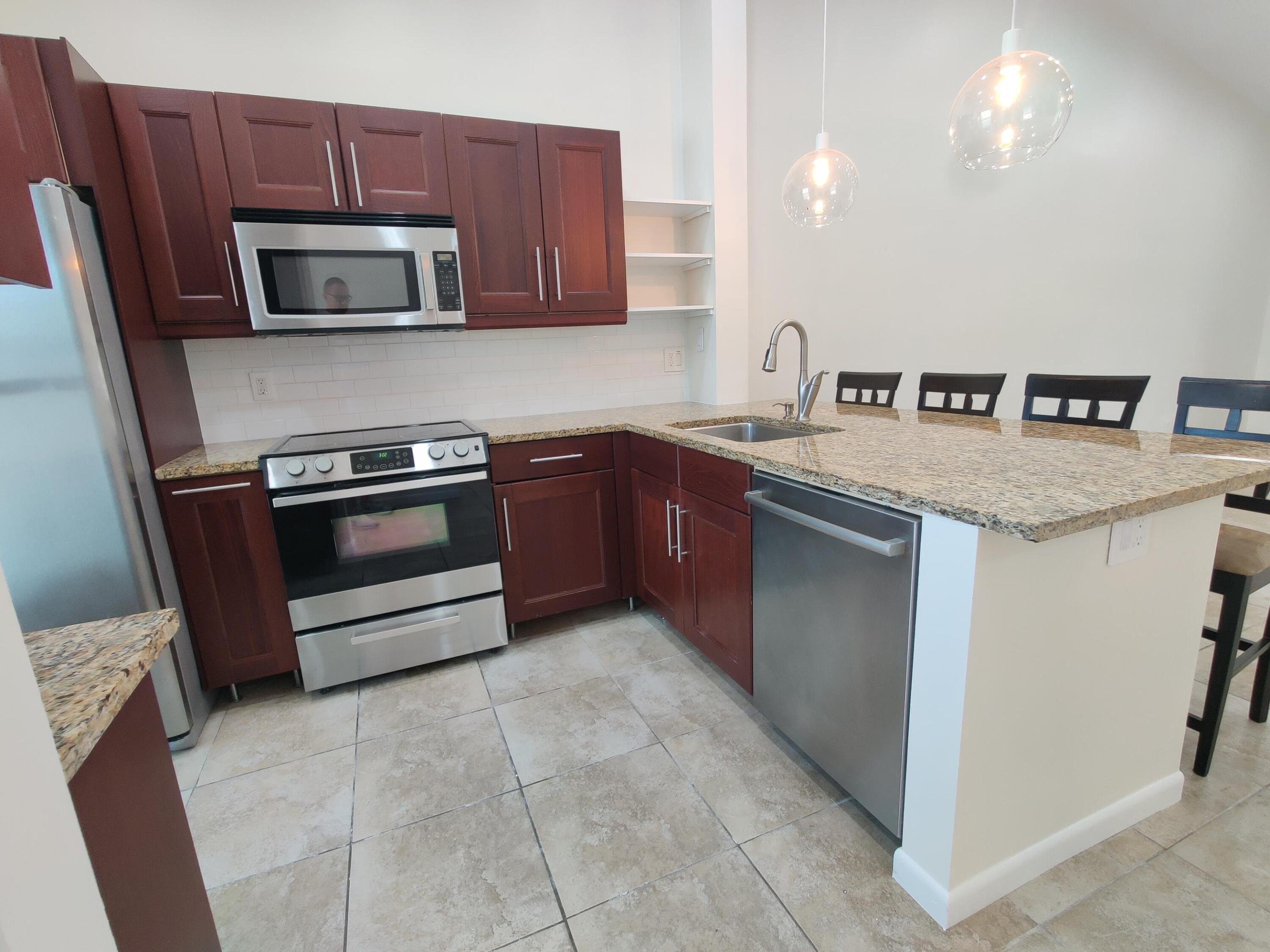 Kitchen featuring stained wood cabinetry, stone-surface countertops, stainless steel appliances, tile flooring, and pendant lighting