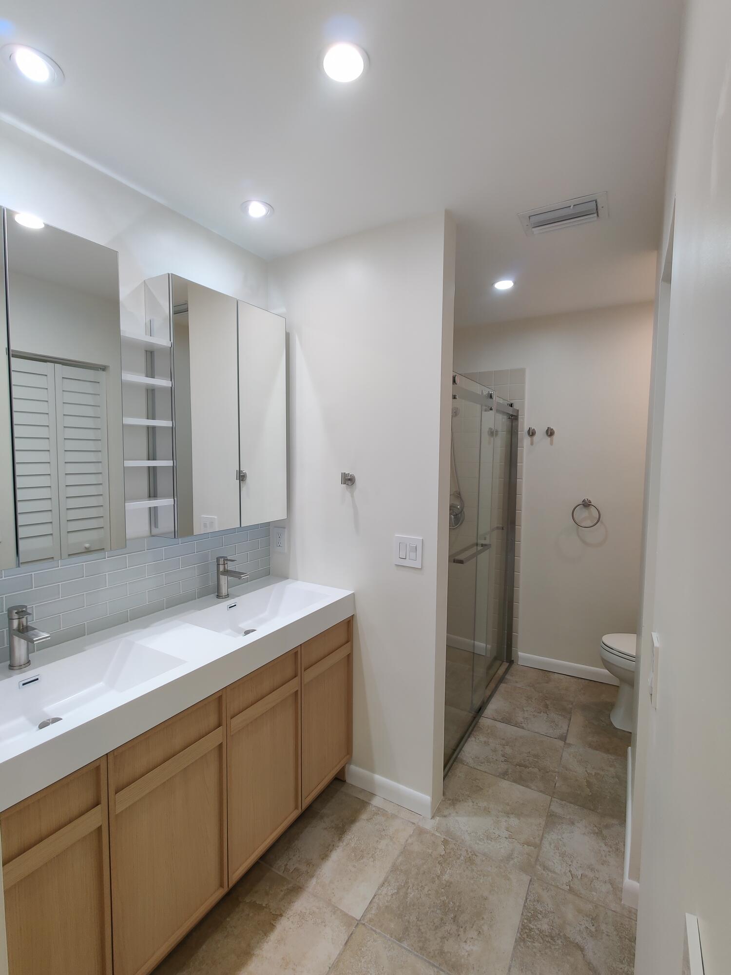 Double vanity with integrated sinks, wood-finish cabinetry, mirrored medicine cabinets, tiled flooring, and a glass-enclosed shower