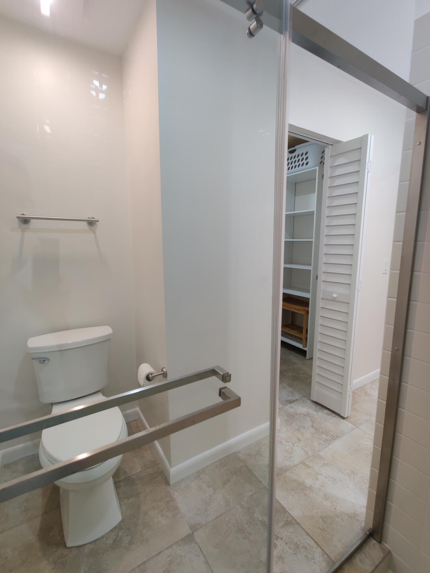 Bathroom featuring a glass-enclosed shower, light-toned stone-finish tile flooring, a dual-tier metal towel rack, and an entry to a storage space with white louvered doors