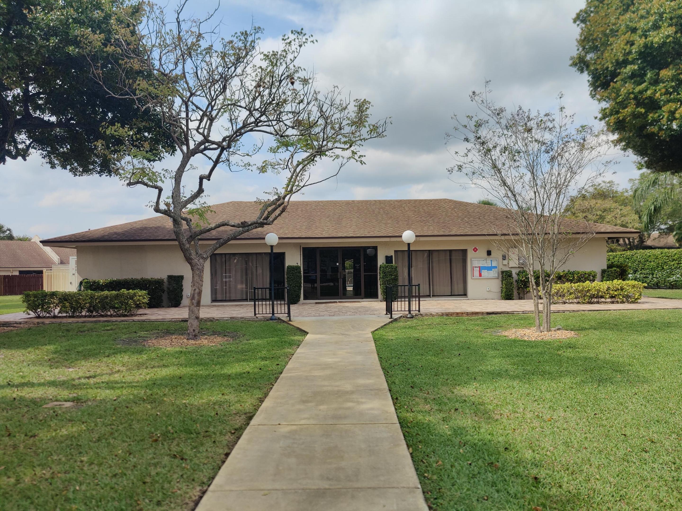 Single-story exterior featuring a concrete walkway, paved patio, mature trees, manicured landscaping, and globe-style pedestal lighting