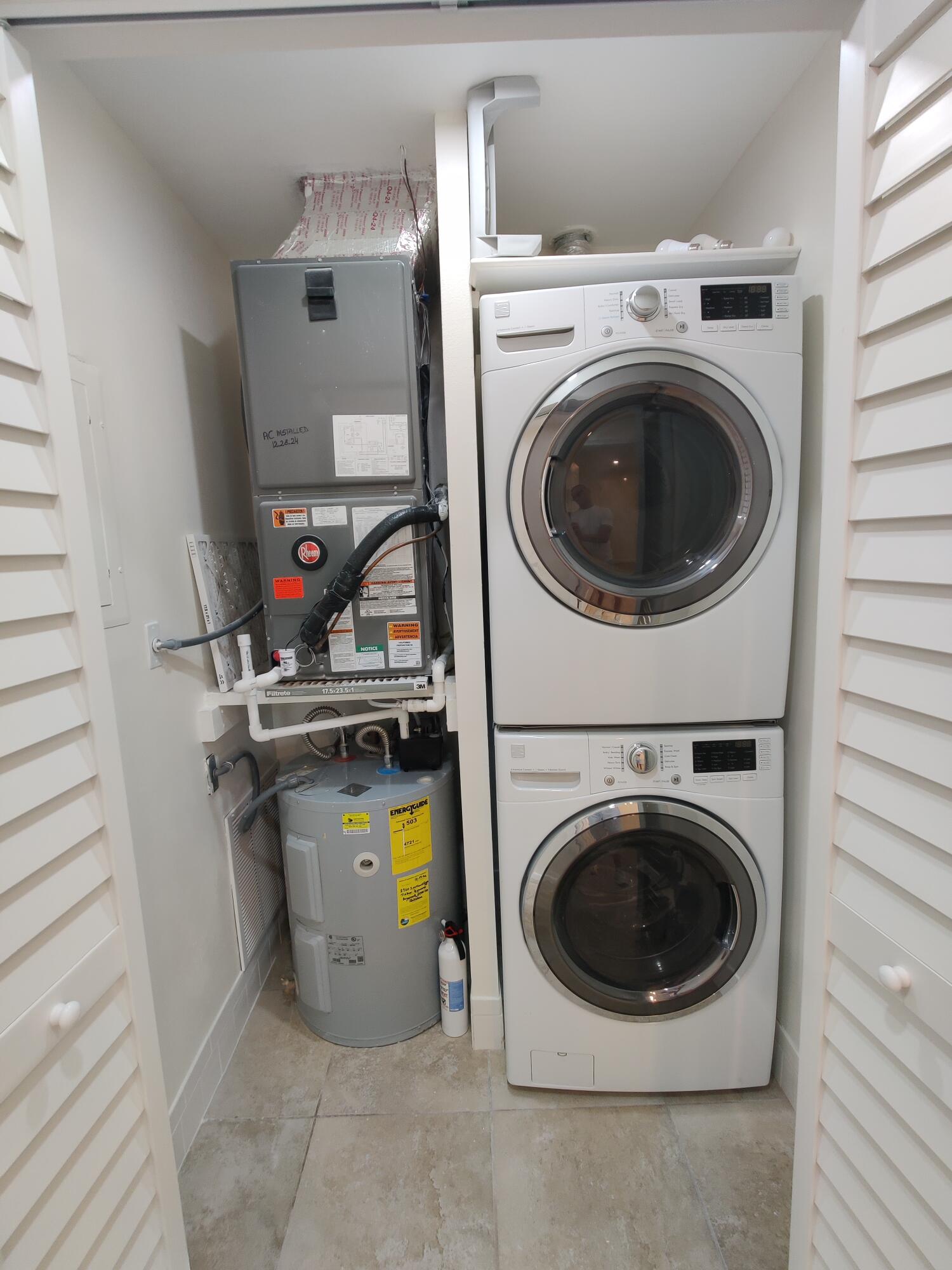 Utility closet featuring a stacked washer and dryer, an integrated water heater, and a climate control air handler unit, enclosed by louvered bi-fold doors with tiled flooring