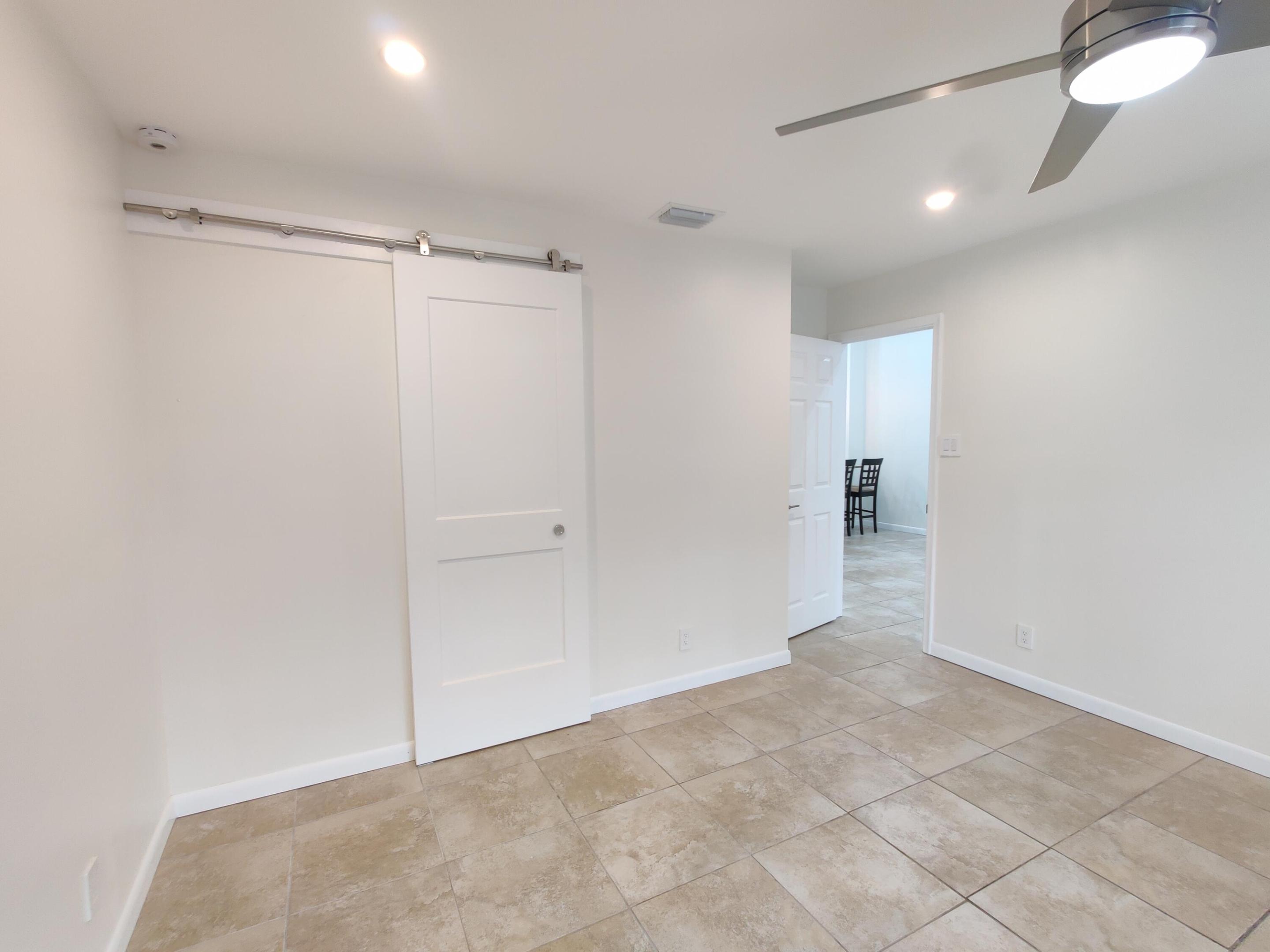 Interior featuring neutral tile flooring, a white two-panel sliding barn door with stainless steel hardware, and a ceiling fan with integrated lighting