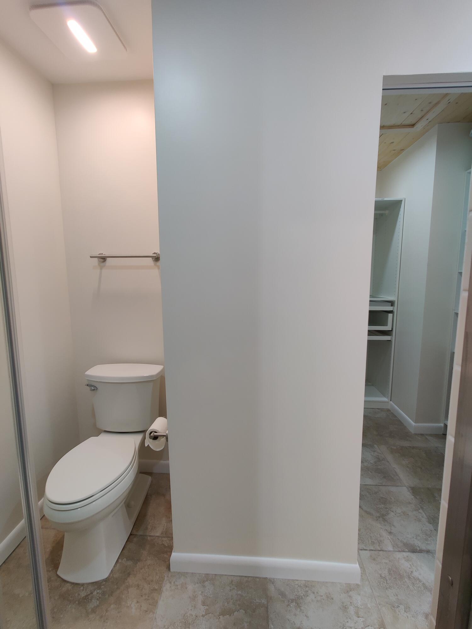 Ceramic tile flooring, white porcelain toilet, wall-mounted metal towel bar, and built-in closet storage featuring shelving and a hanging rod