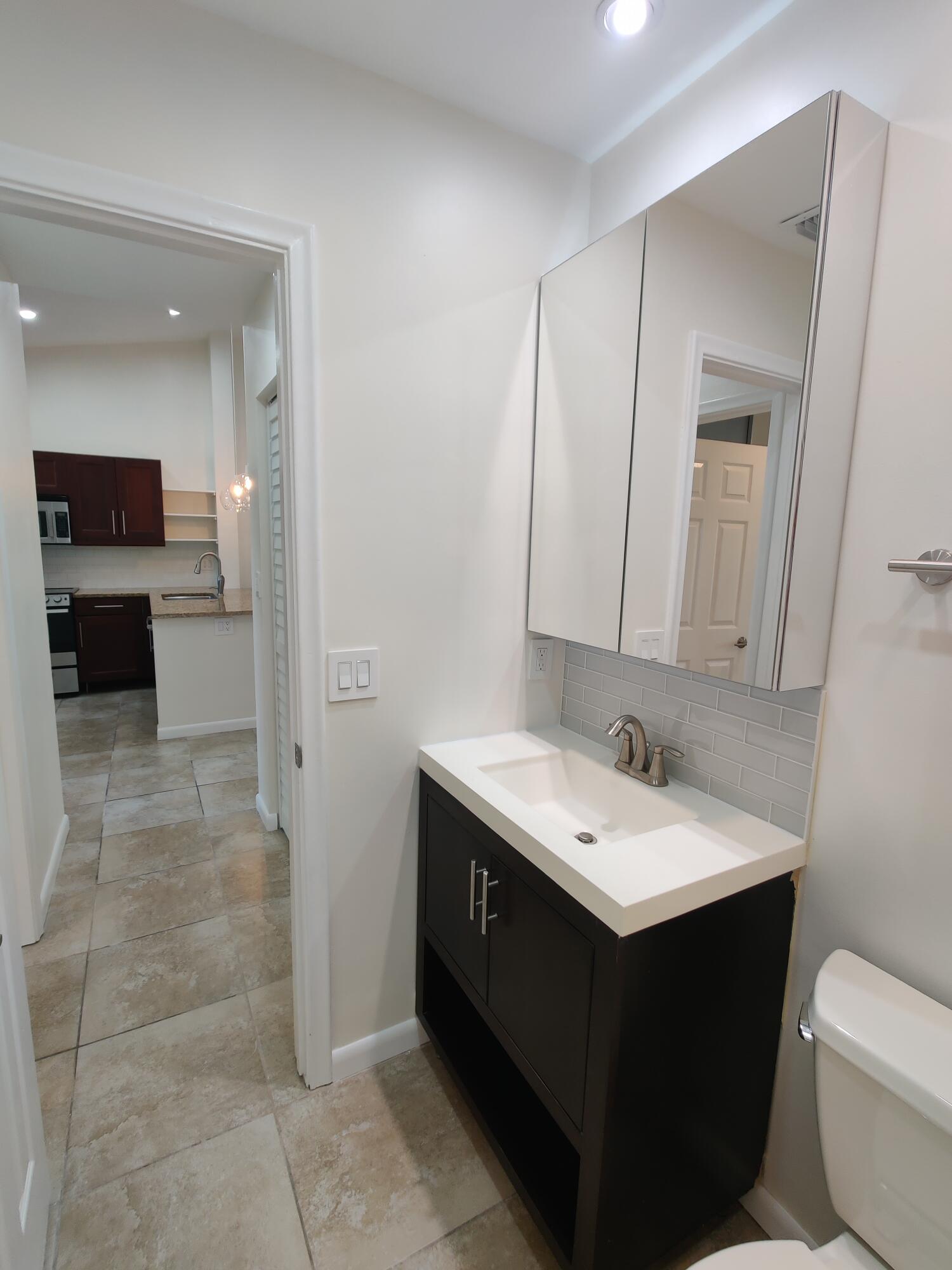 Dark wood vanity featuring a rectangular integrated sink, neutral stone-finish countertop, mirrored medicine cabinet, and tile backsplash