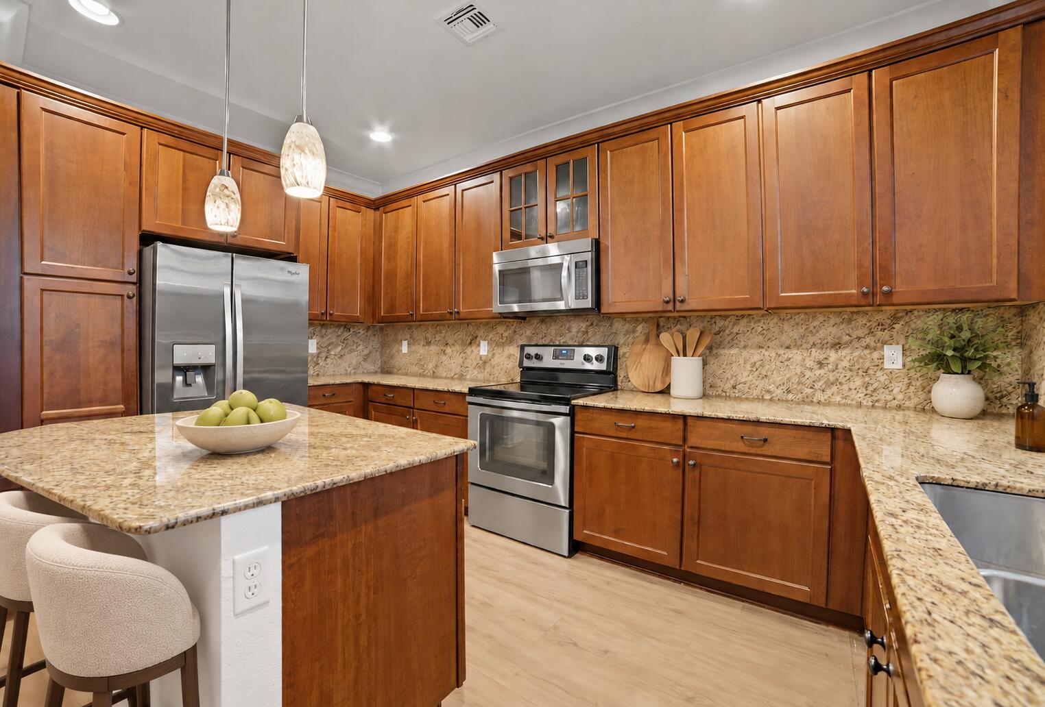 Kitchen featuring medium-toned wood cabinetry, stone countertops, stainless steel appliances, a central island, and glass pendant lighting