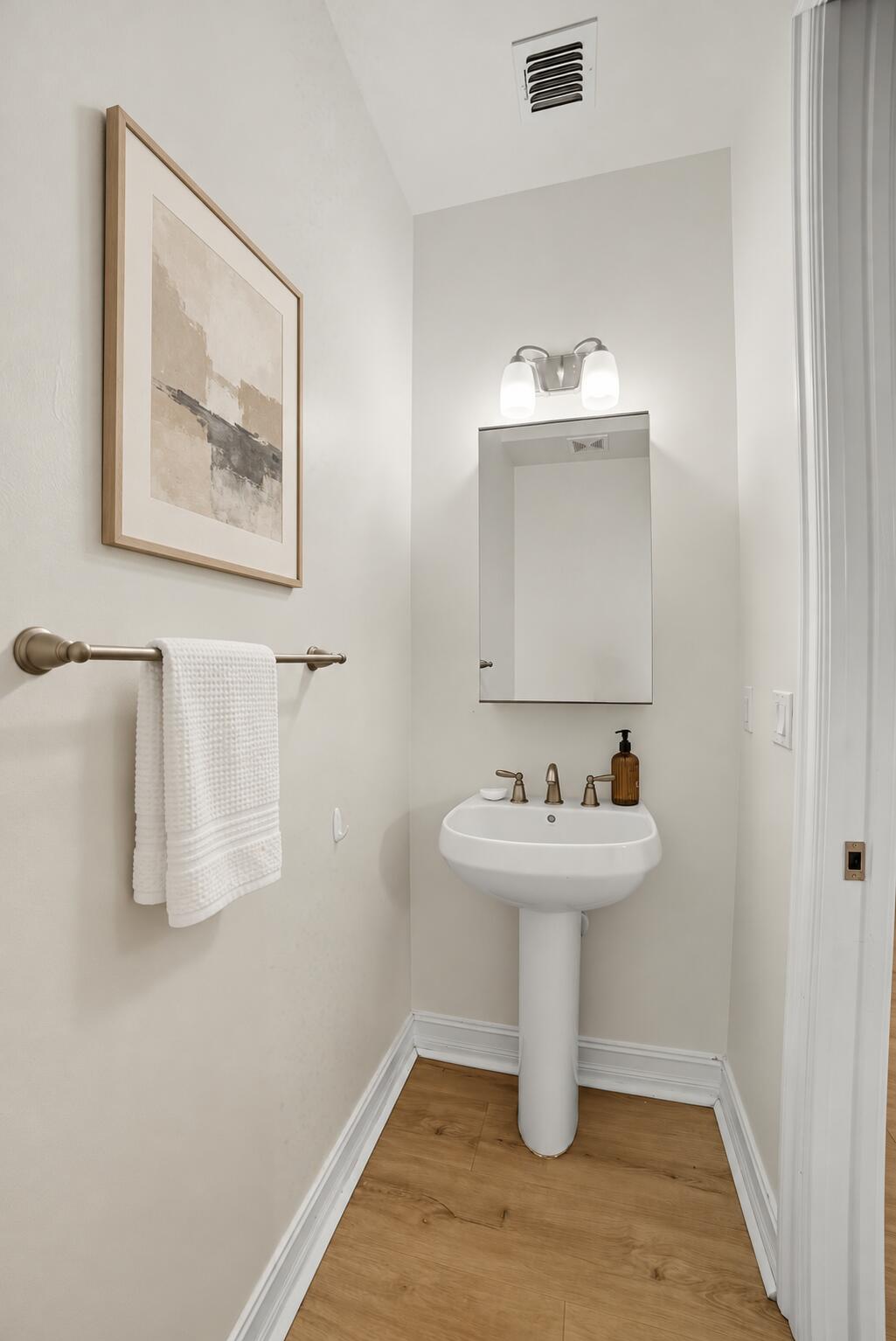 White pedestal sink with a metallic widespread faucet, recessed medicine cabinet, dual-globe wall-mounted light fixture, and wood-finish flooring