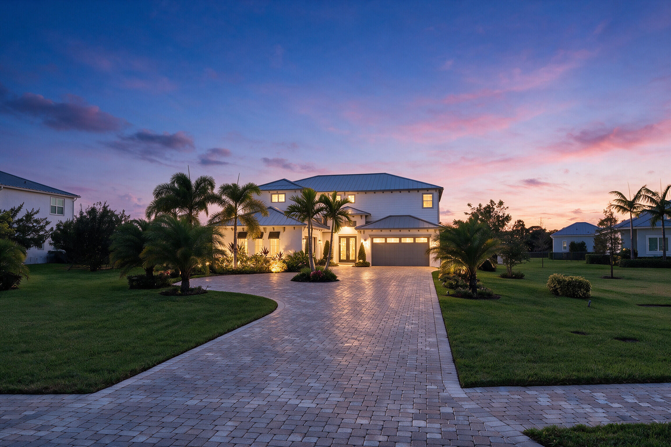 Two-story exterior featuring light-toned masonry, a multi-car garage, and an expansive paved driveway surrounded by mature palm trees and landscaped lawns