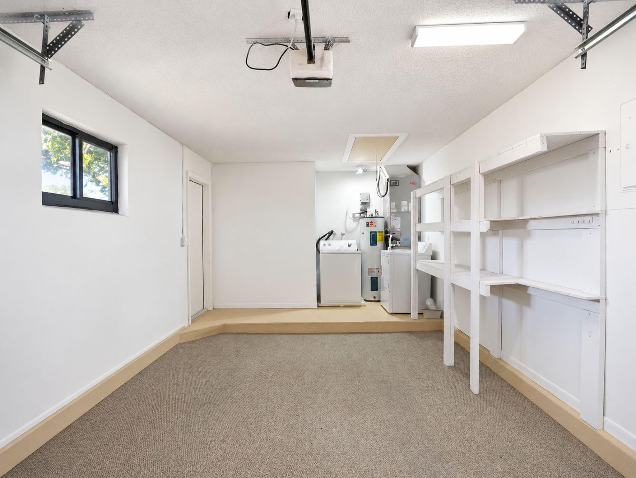 Enclosed garage space featuring carpeted flooring, a raised utility platform, built-in wall shelving, a single-pane window, and an integrated water heater