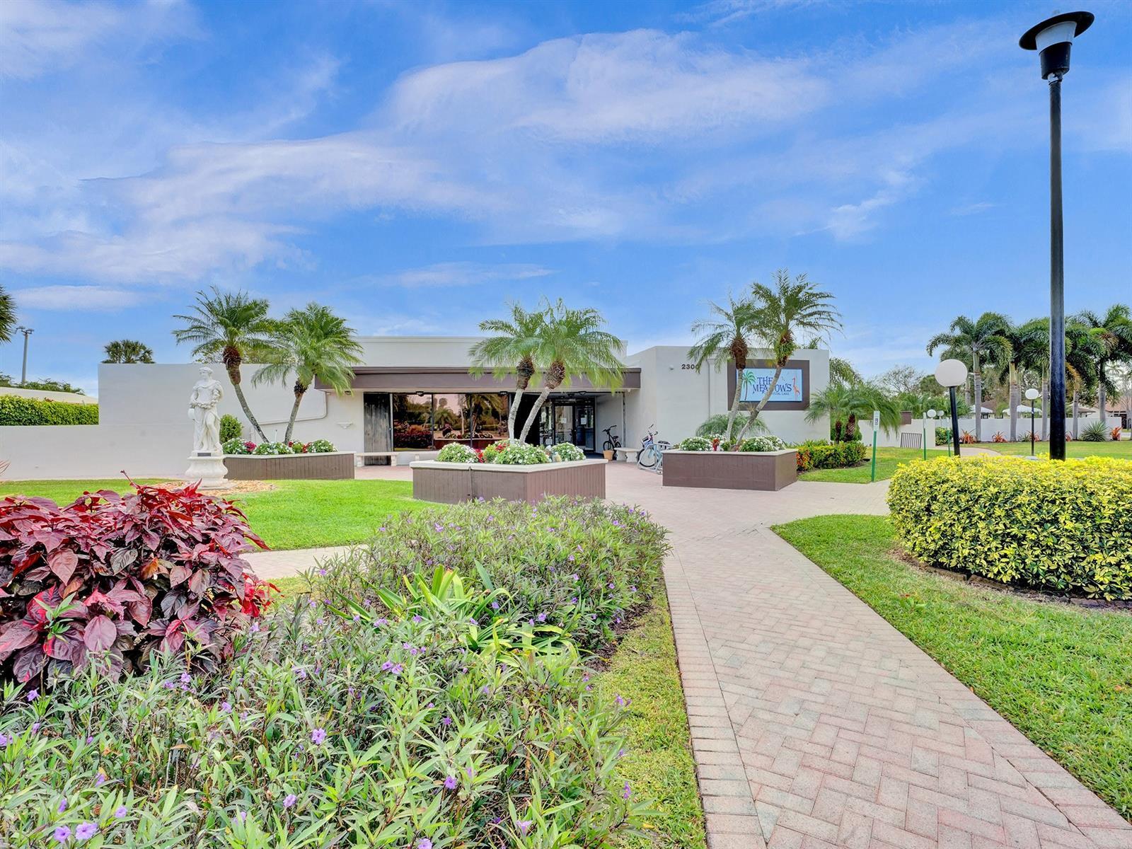 Single-story clubhouse exterior featuring stucco siding, paved walkways, manicured landscaping with mature palm trees, and decorative masonry planters