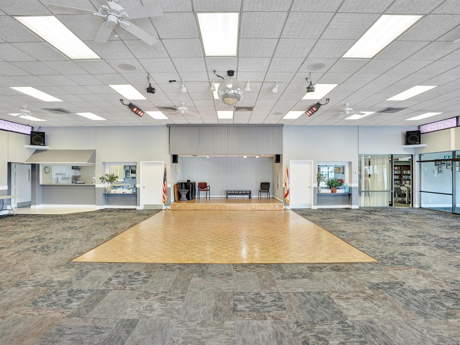 Open-concept event space featuring a central raised stage, a dedicated hardwood-finish dance floor, acoustic tile drop ceilings with recessed lighting, and integrated service counters